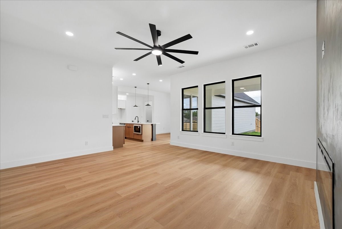 Vacant living room with light wood floors, a large ceiling fan, and three tall windows beside the open kitchen.
