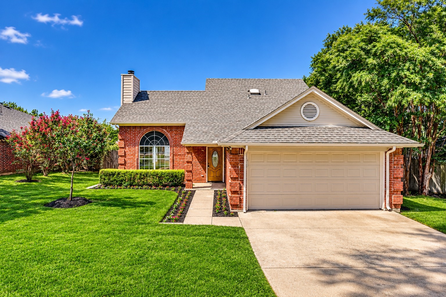 Red-brick home with an arched window, attached garage, flower beds, and broad green lawn.