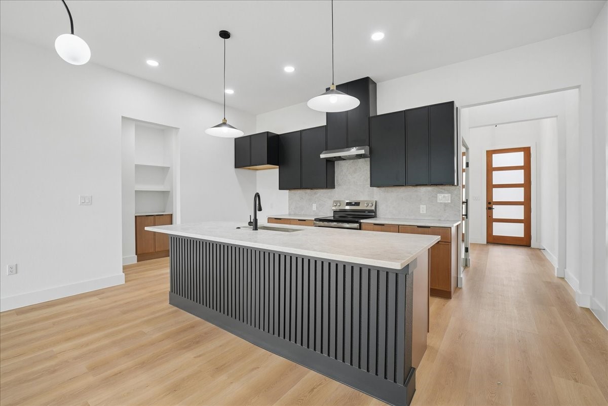Vacant modern kitchen with a large island, dark cabinetry, stainless steel appliances, and light wood floors.
