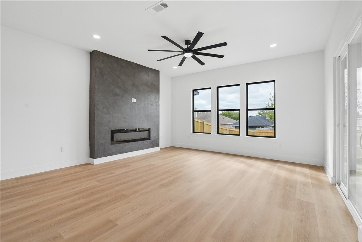 Empty living room with a dark fireplace wall, light wood floors, and three large windows.
