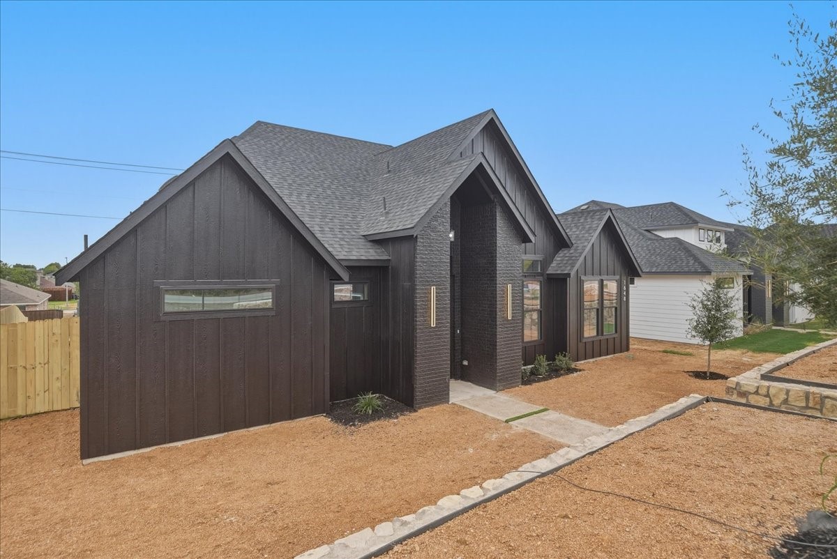 Modern home with dark vertical siding and brick accents, viewed from the front corner beside a gravel yard.