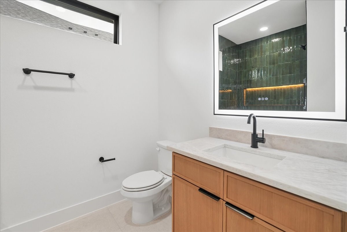 Modern bathroom with a light wood vanity, illuminated mirror, dark green tile, and black fixtures.