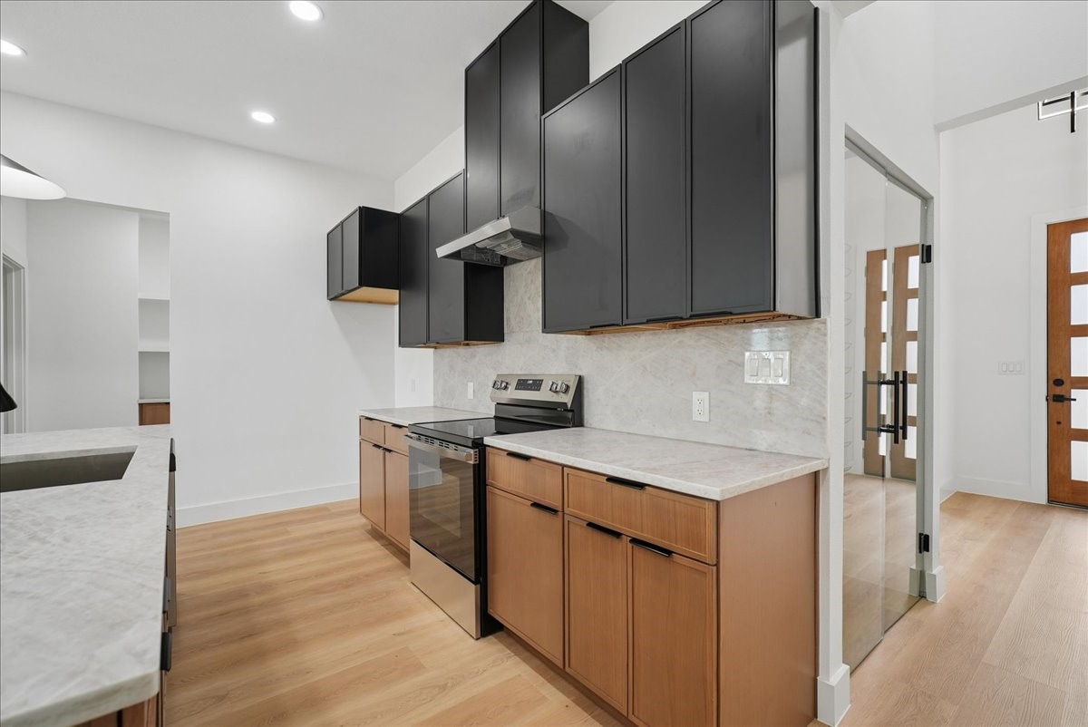 Modern kitchen with charcoal cabinets, warm wood lower cabinets, pale stone counters, and a stainless steel range.