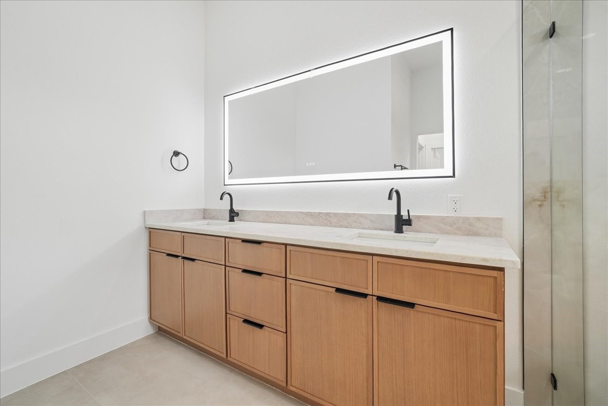 Bathroom vanity with light wood cabinets, dual black faucets, an illuminated mirror, and a glass shower.