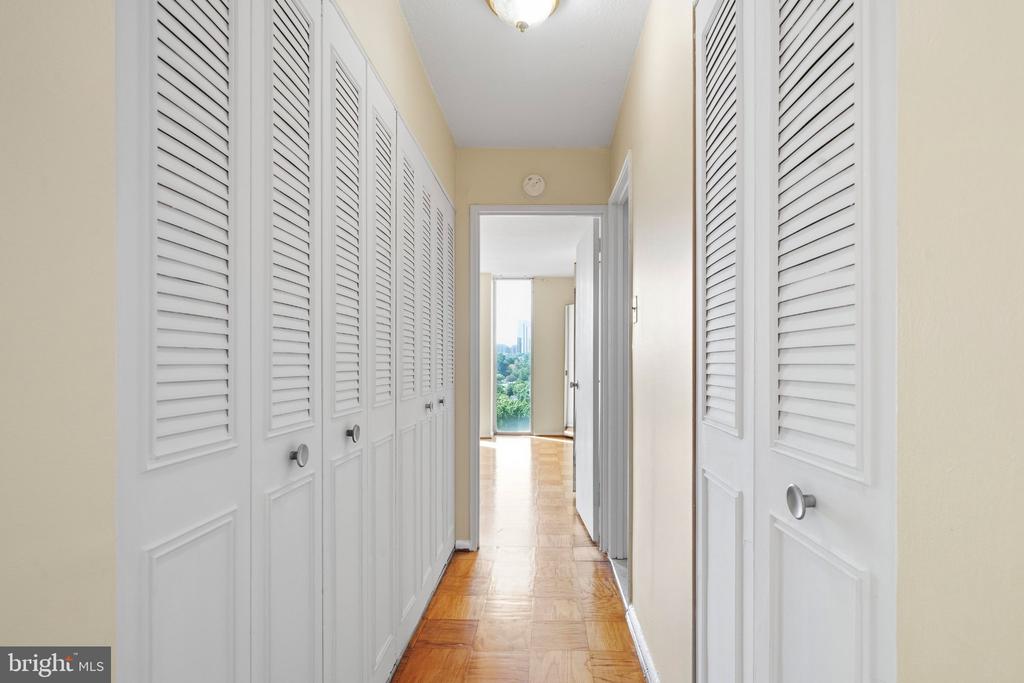 Long hallway with white louvered closet doors, parquet flooring, and greenery visible through a distant window.