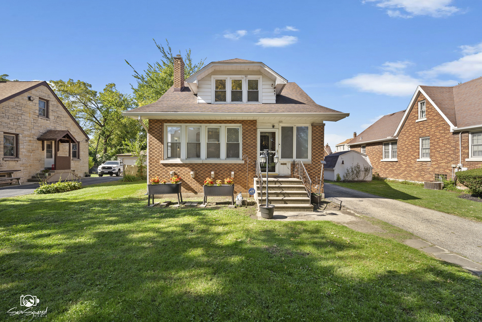 Brick house with a white dormer, flower planters, and front steps beside a green lawn.