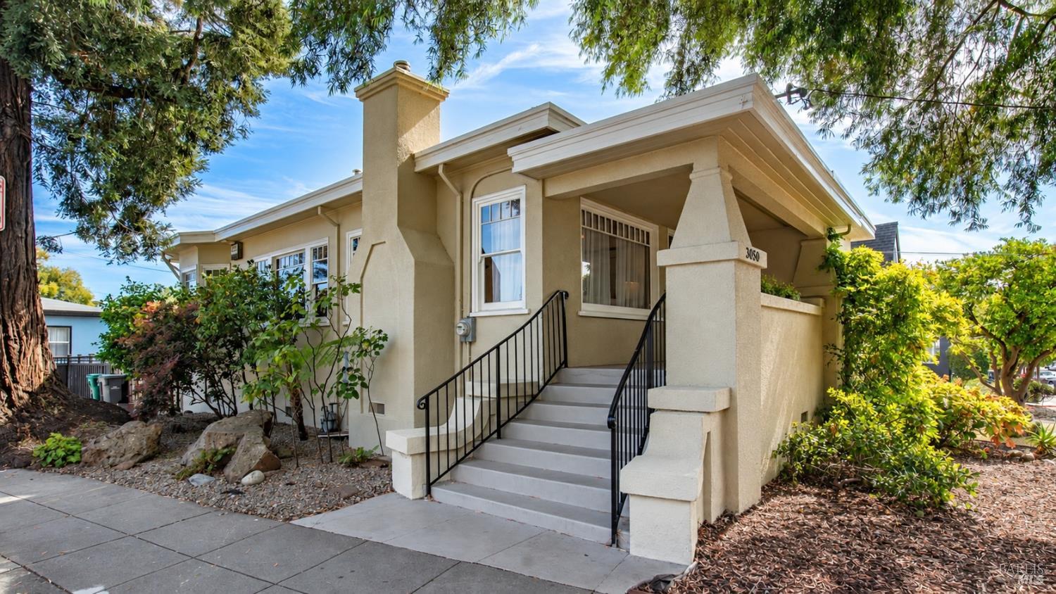 Beige stucco home with a covered porch, broad front steps, and black railings, framed by mature trees.