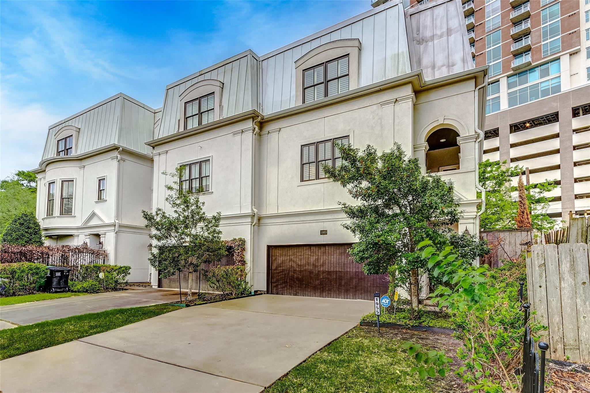 White multi-story home with a mansard-style roof, arched entry, and broad driveway beside a high-rise.