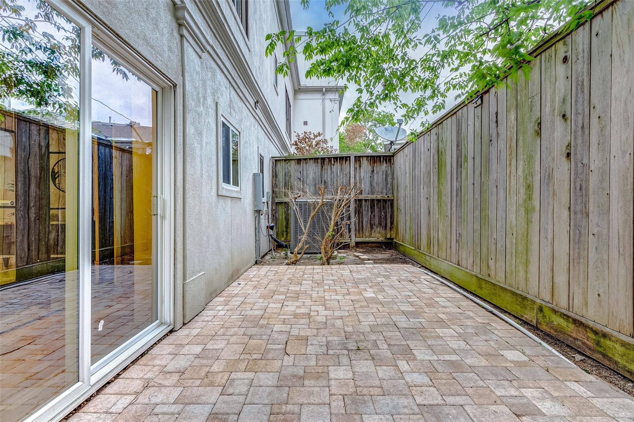 Paved side patio enclosed by tall wooden fencing, with a sliding glass door and bare shrub.