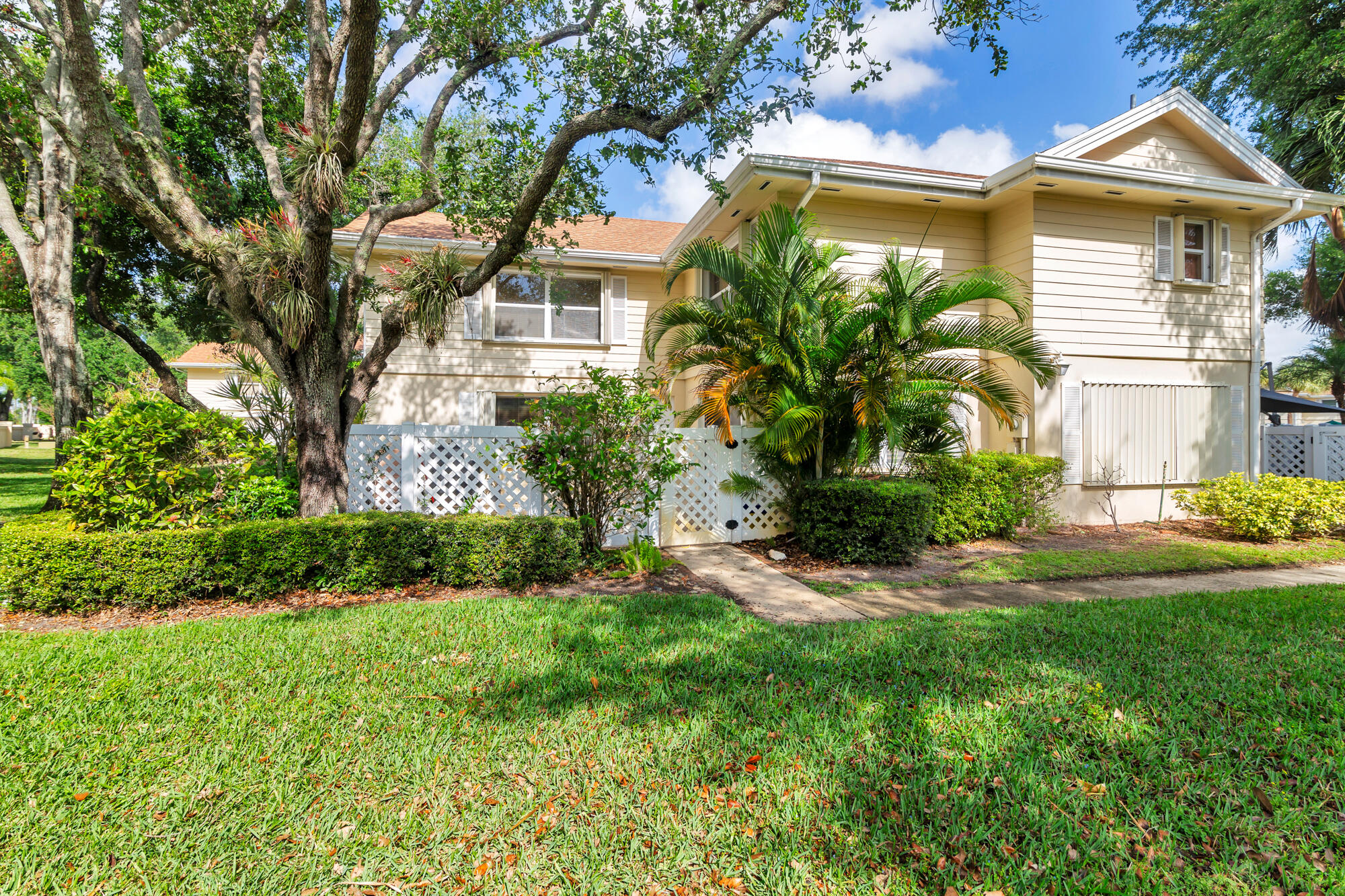 Two-story beige home framed by mature trees and tropical palms, with a green lawn and hedges.