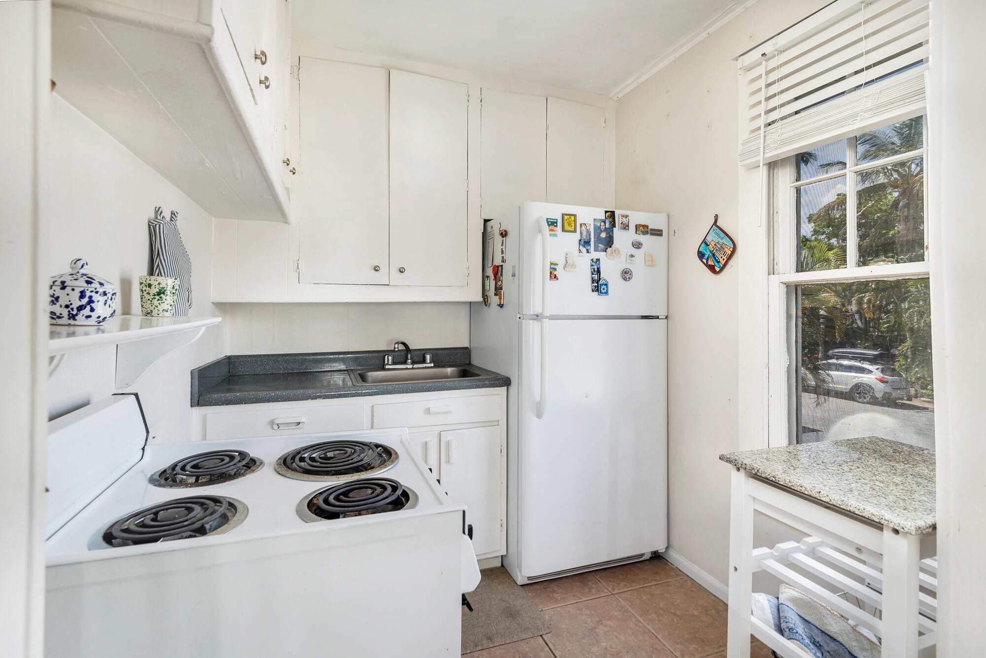 Compact kitchen with white cabinets, coil range, refrigerator, and a window overlooking trees.