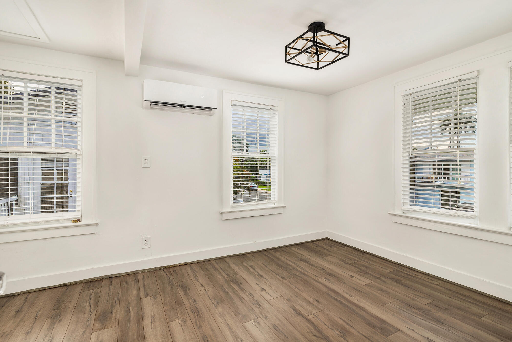 Empty room with wood-look flooring, three blinded windows, and a wall-mounted air conditioner.