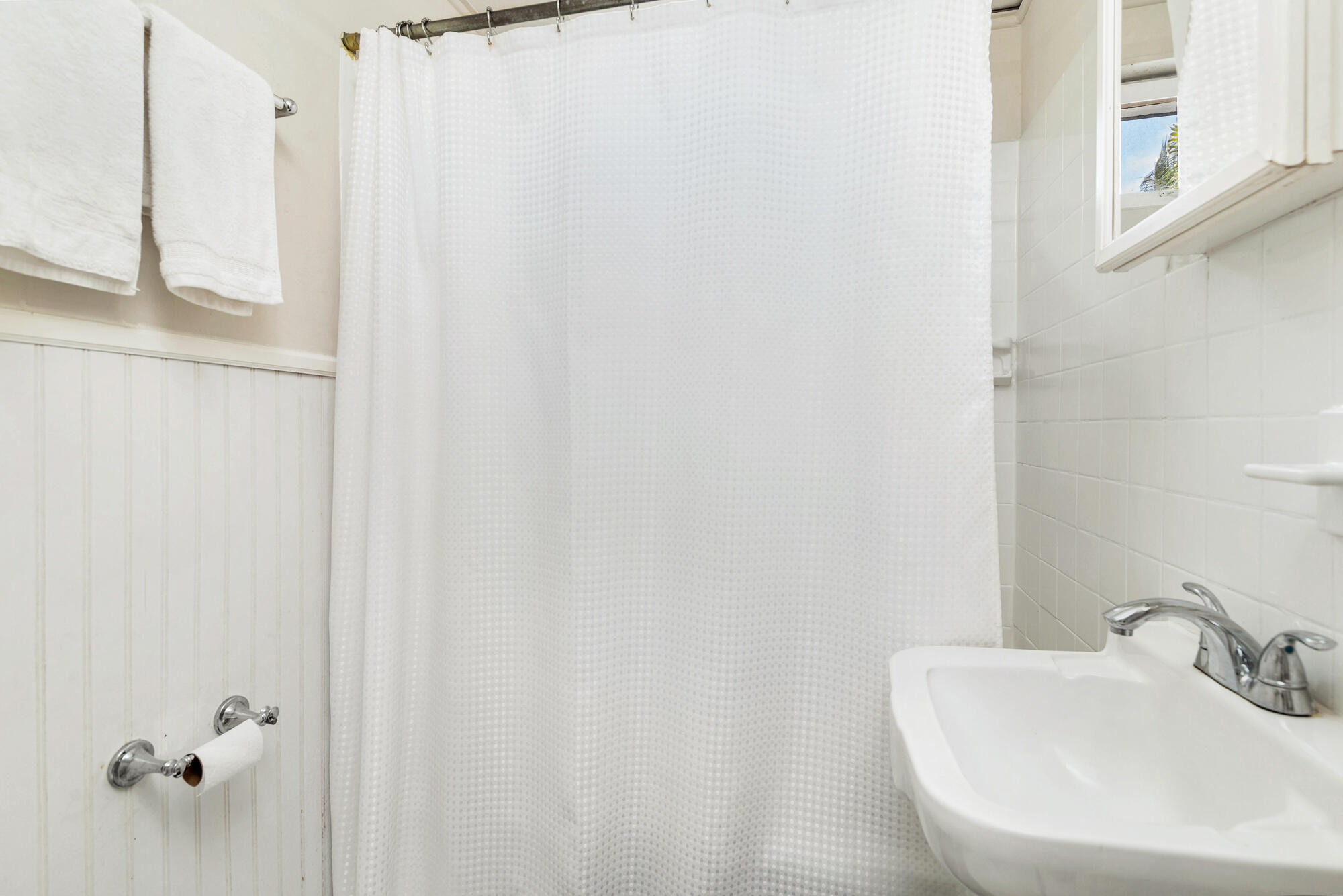 White bathroom with a shower curtain, tiled walls, and a wall-mounted sink beneath a window.