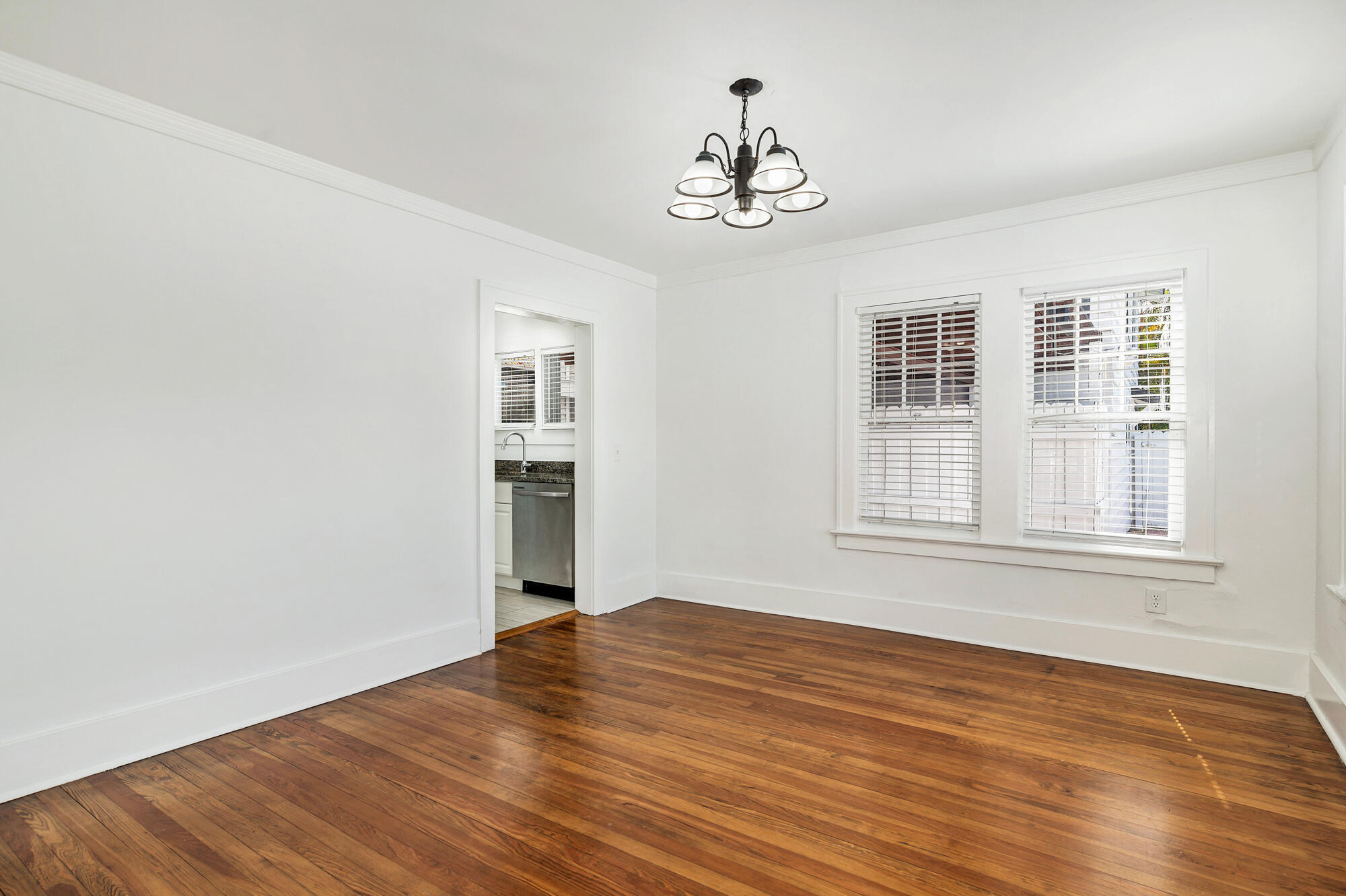 Empty room with hardwood floors, a chandelier, two blinded windows, and a kitchen doorway.