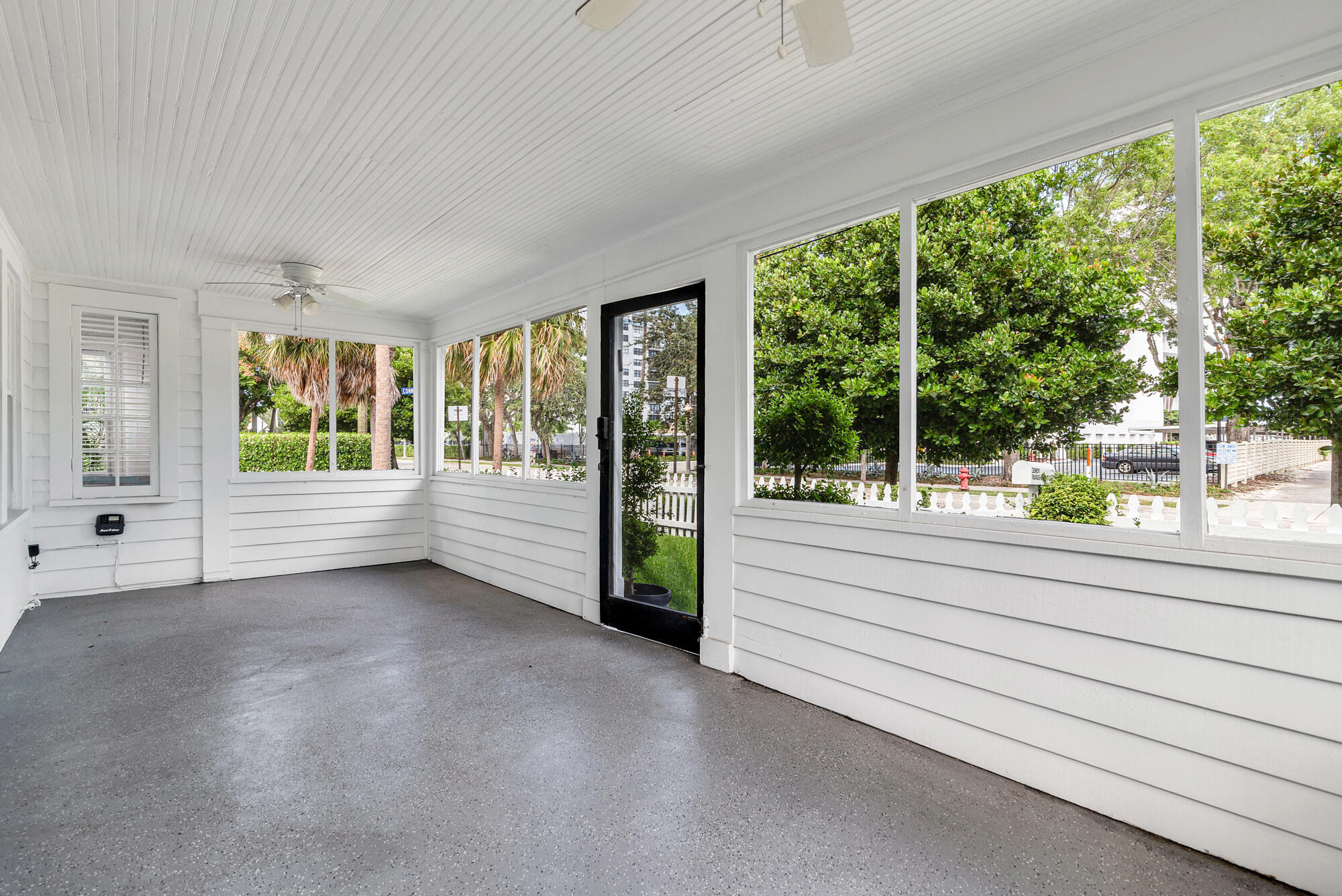 Enclosed porch with white walls, a ceiling fan, gray floor, and large windows overlooking trees.