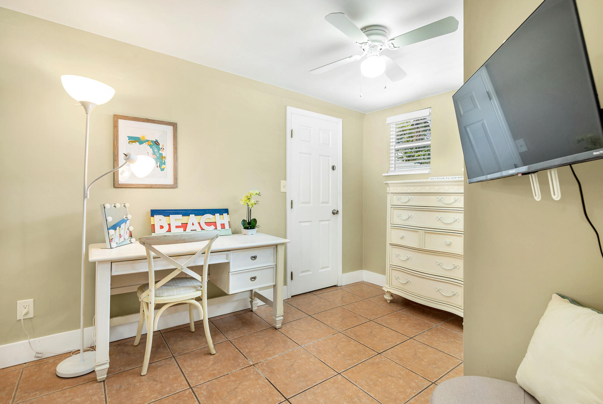 Furnished bedroom with a white desk, ornate dresser, terracotta tile floor, and wall-mounted television.