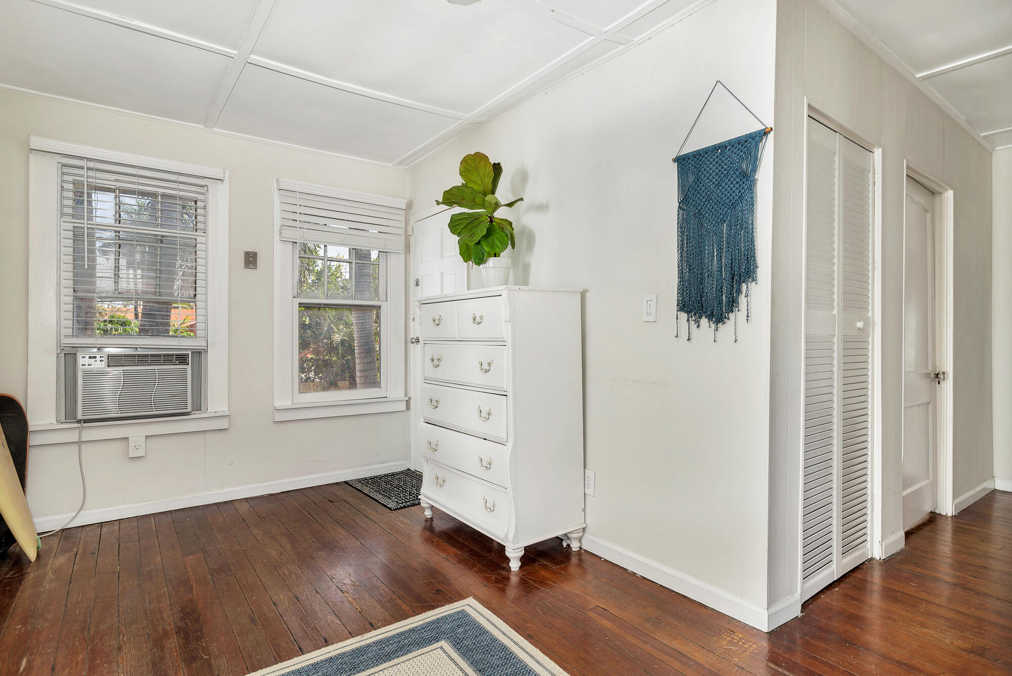 Bedroom with dark hardwood floors, a white dresser and plant beside two windows with blinds.