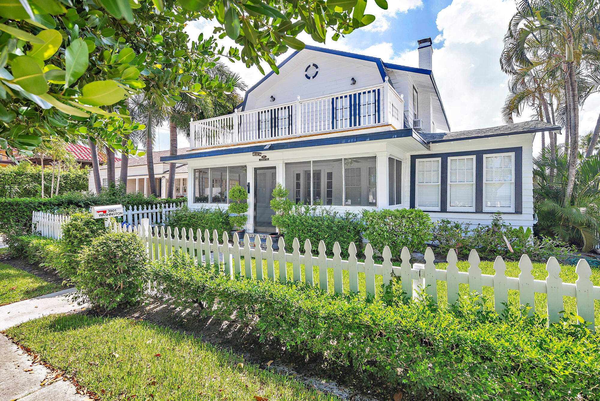 White two-story house with navy trim, a balcony, screened porch, and tropical landscaping behind a picket fence.
