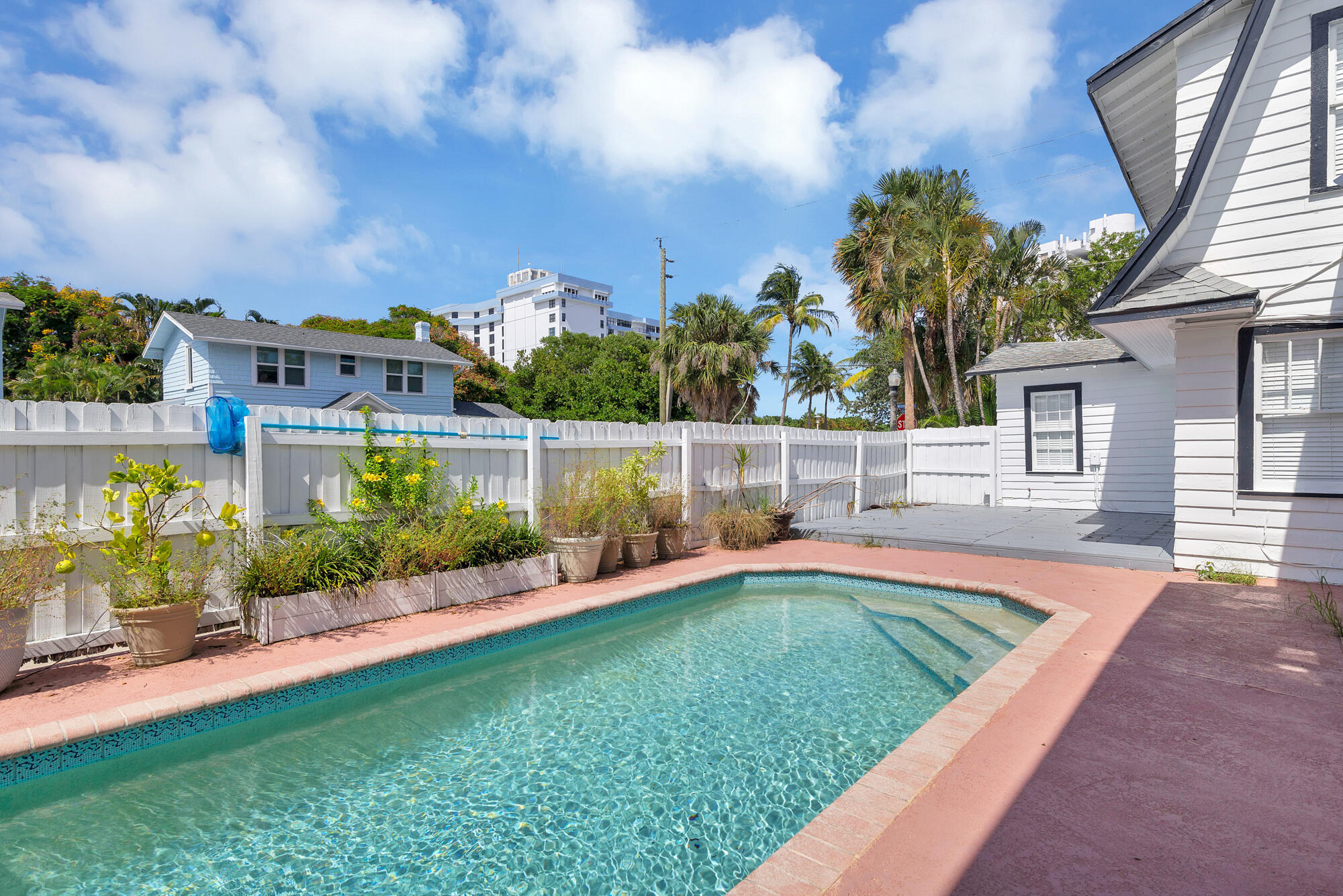 Outdoor swimming pool beside a white-sided house, with a pink patio deck and tropical plants.