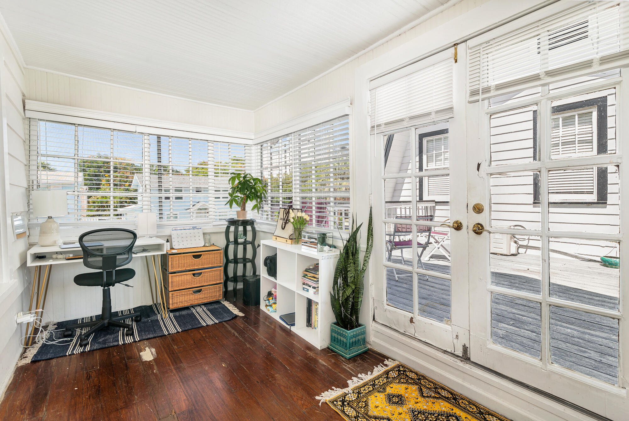 Bright sunroom office with broad windows, French doors to a deck, and dark wood floors.