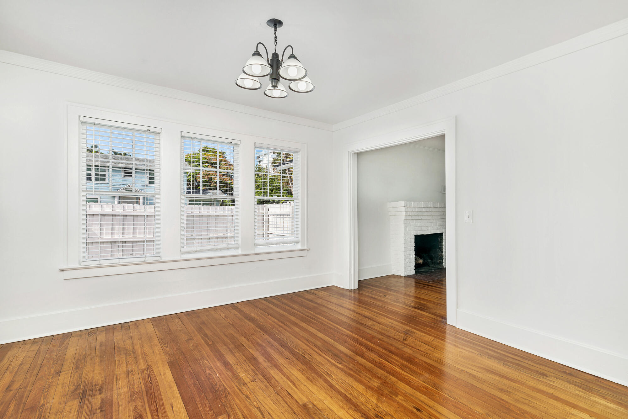 Vacant room with polished hardwood floors, three curtained windows, and a doorway to a white brick fireplace.