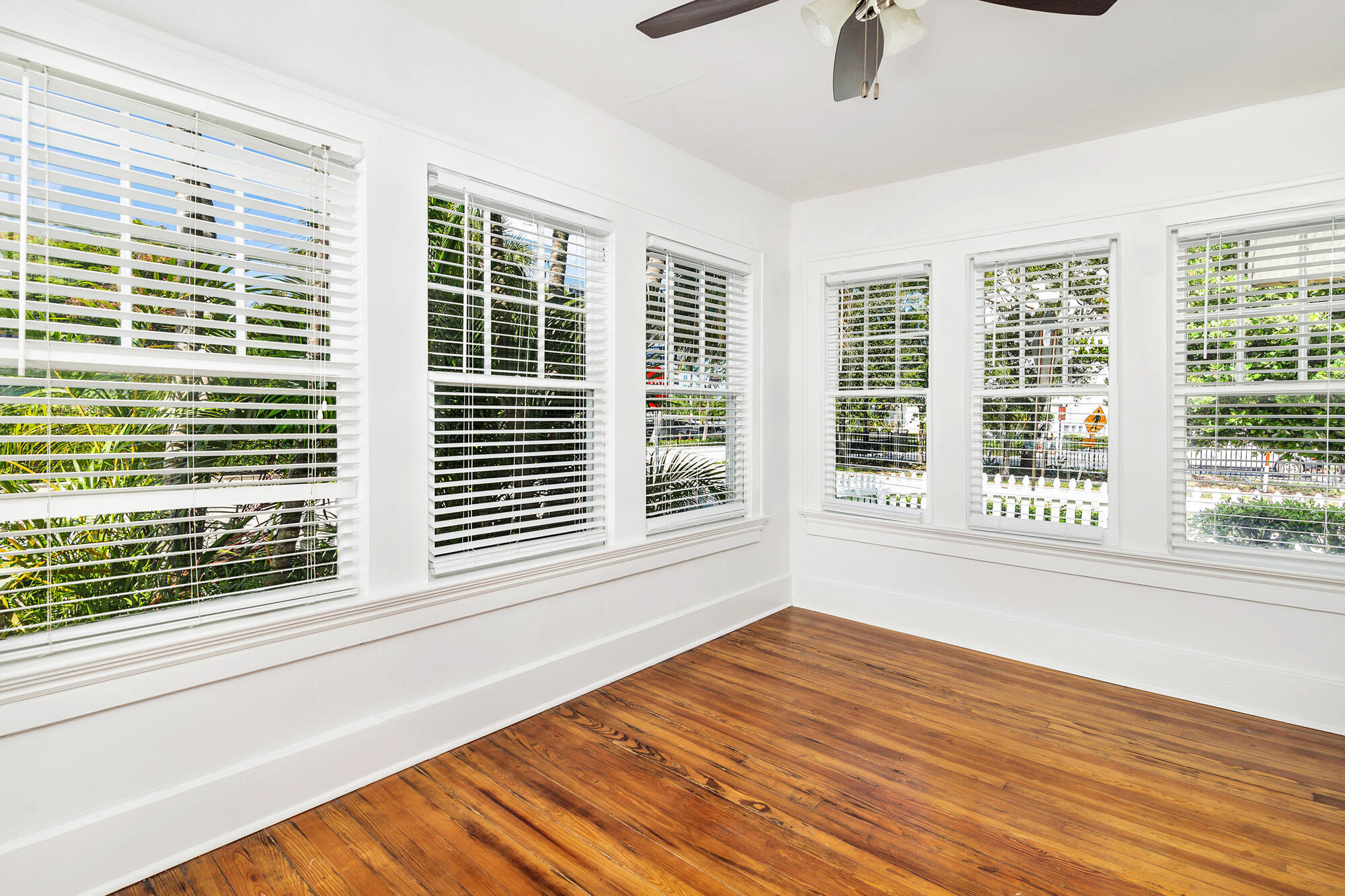 Empty bright room with hardwood floors, white blinds, and windows overlooking greenery.