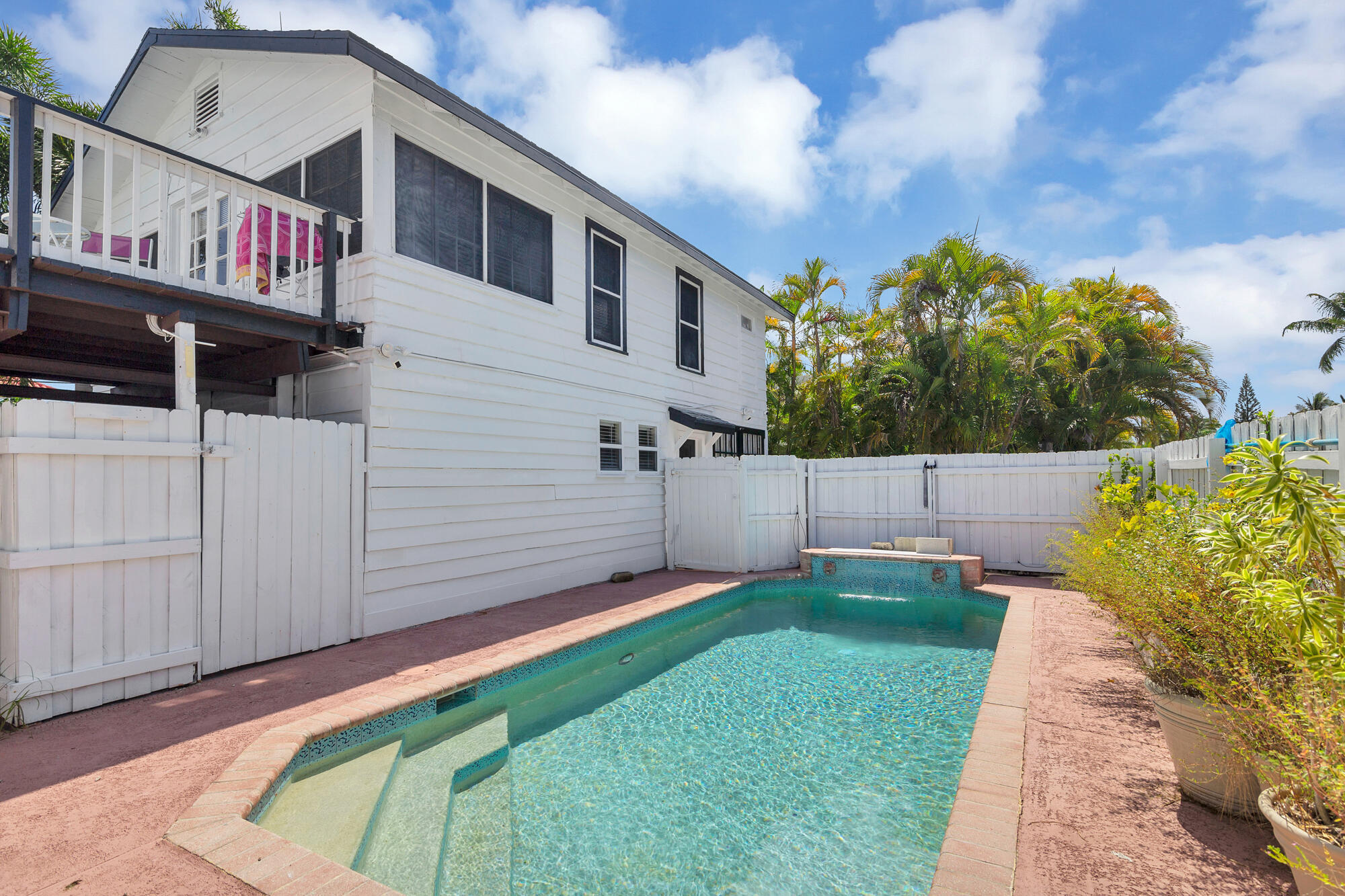 White two-story house beside a turquoise backyard pool, with tropical palms and a fenced patio.