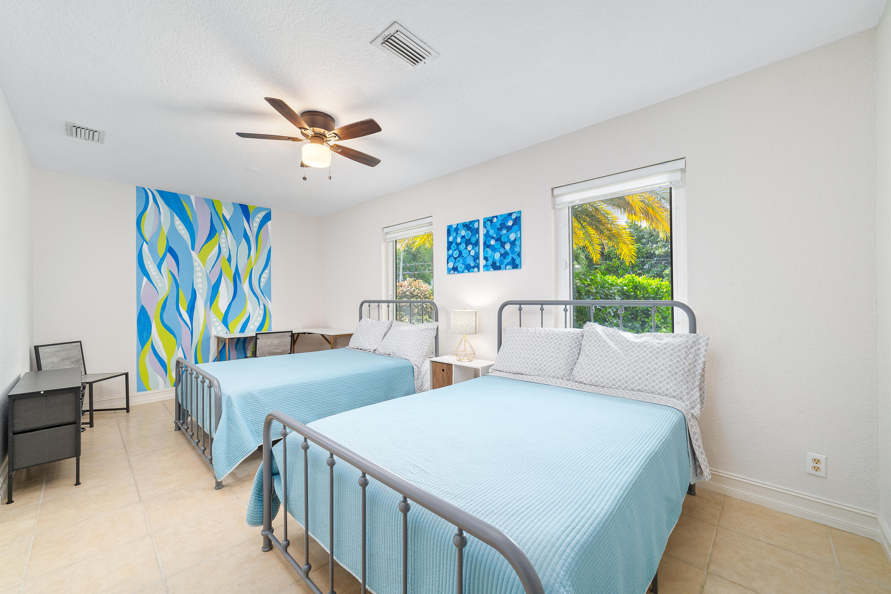 Bedroom with two light blue beds, colorful abstract wall art, and windows overlooking tropical greenery.