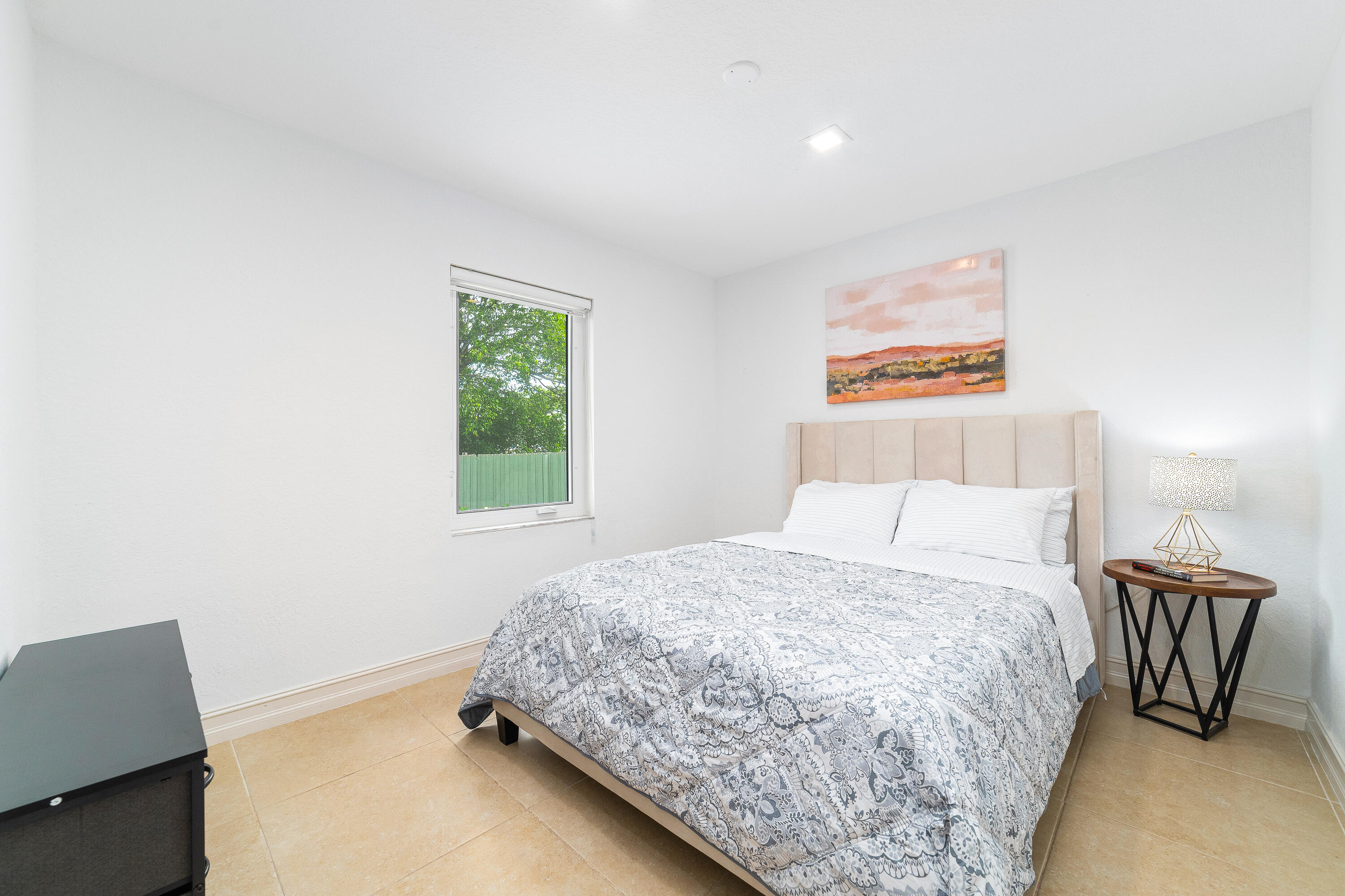 Bedroom with patterned bedding, a pale upholstered headboard, and a window overlooking trees and a green fence.