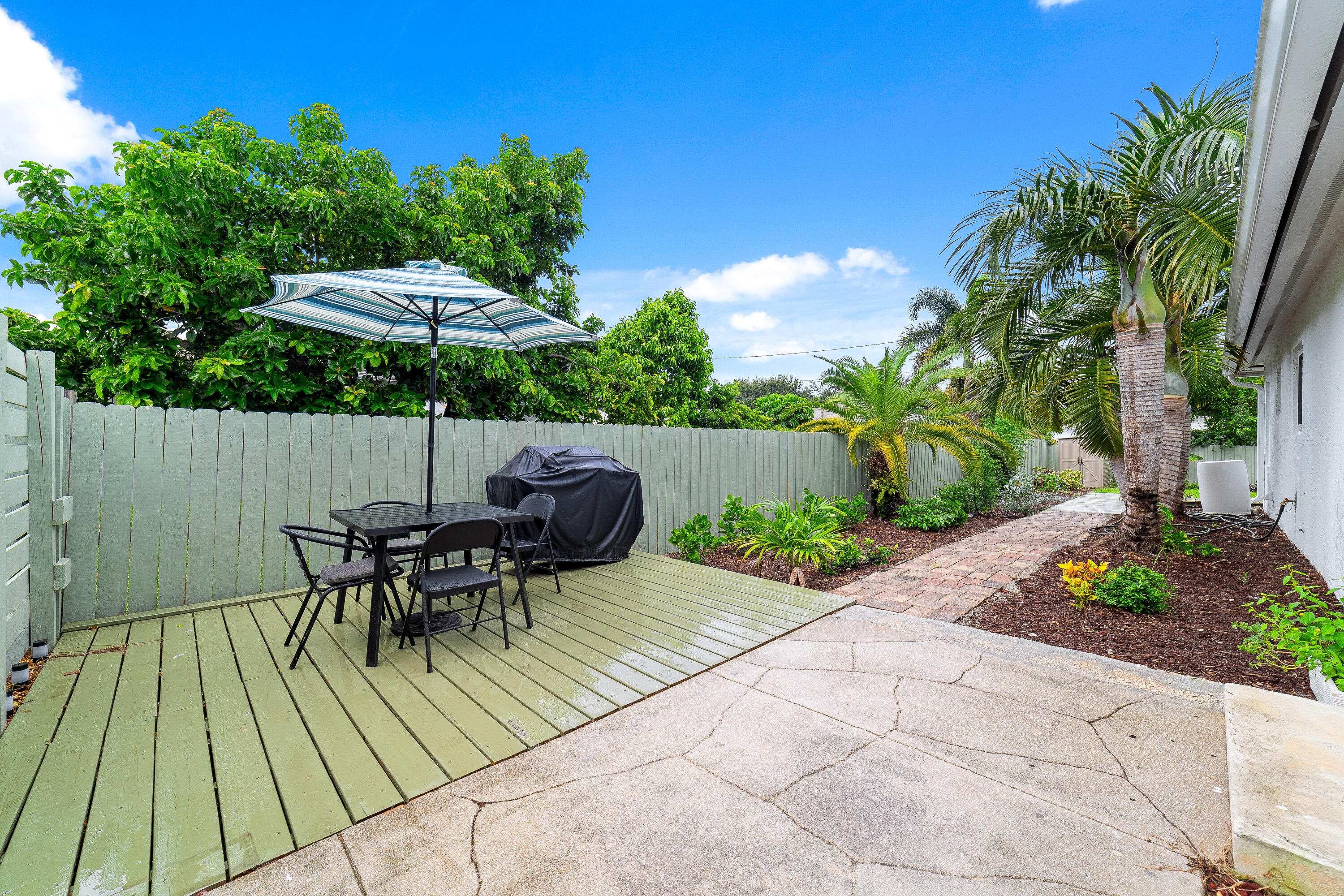 Outdoor patio with a striped umbrella over a dining set beside tropical landscaping and a paver walkway.