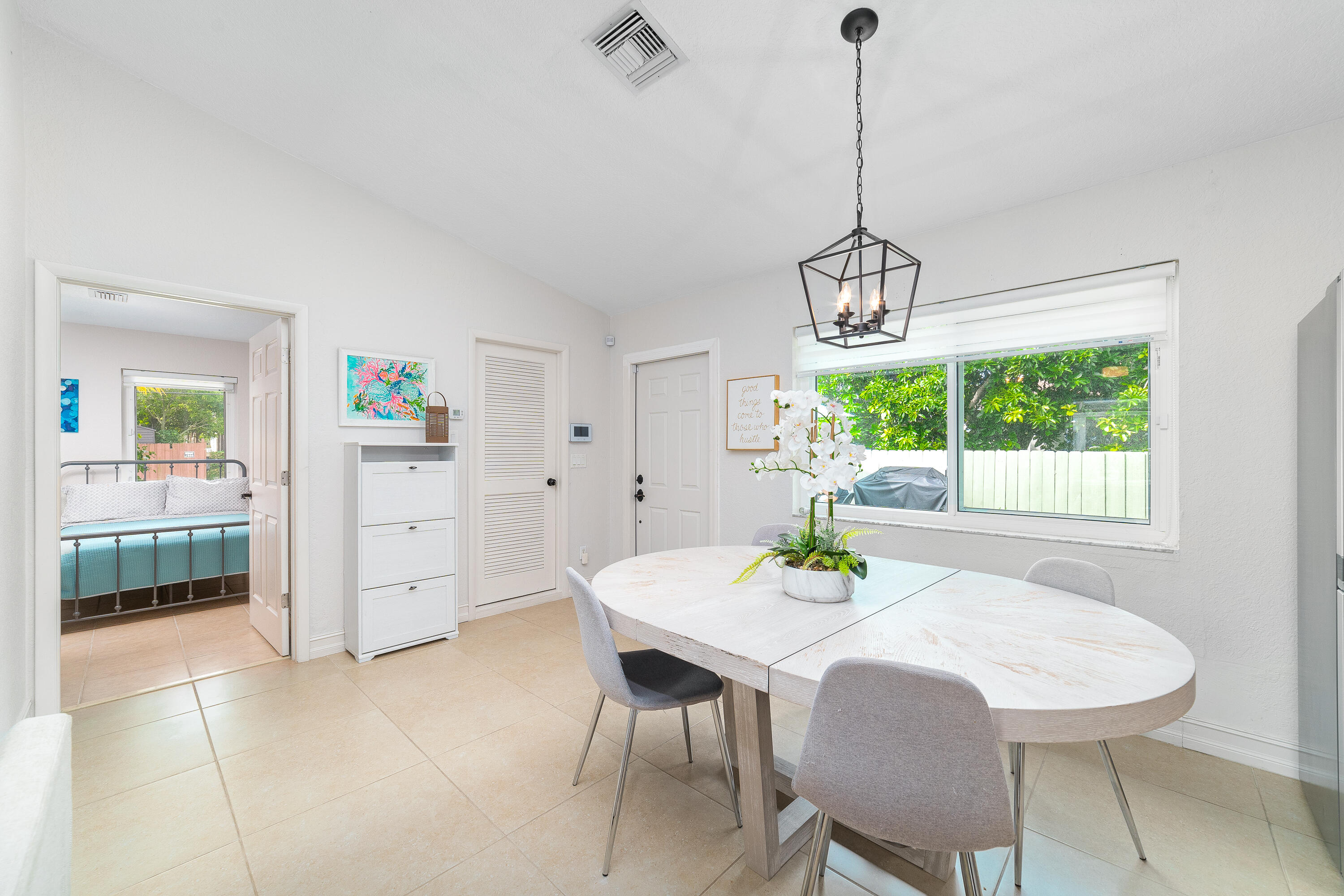 Dining area with a white oval table and chairs beside a large window overlooking a leafy yard.
