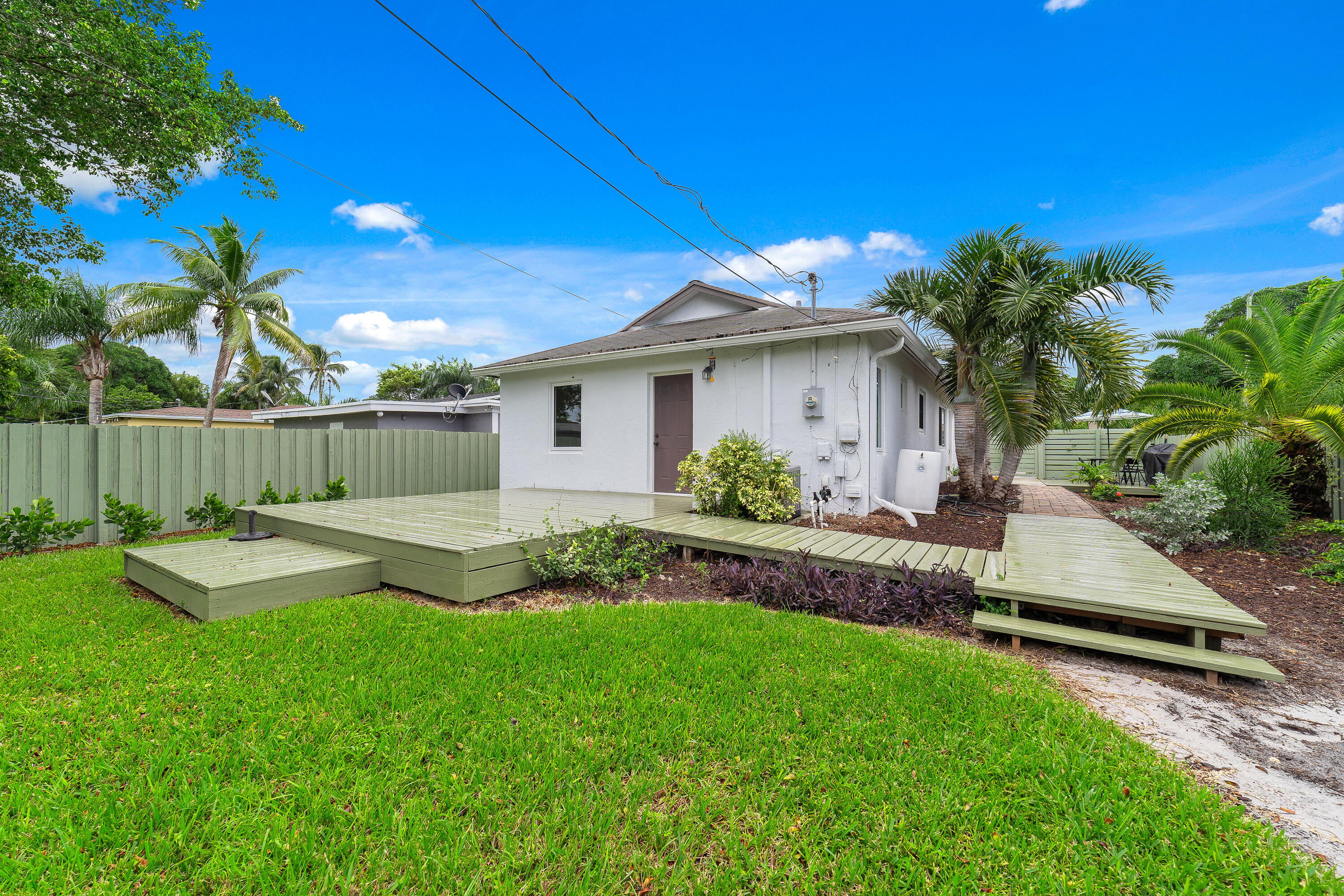 Rear of a white house with a raised deck, green lawn, privacy fence, and palm trees.