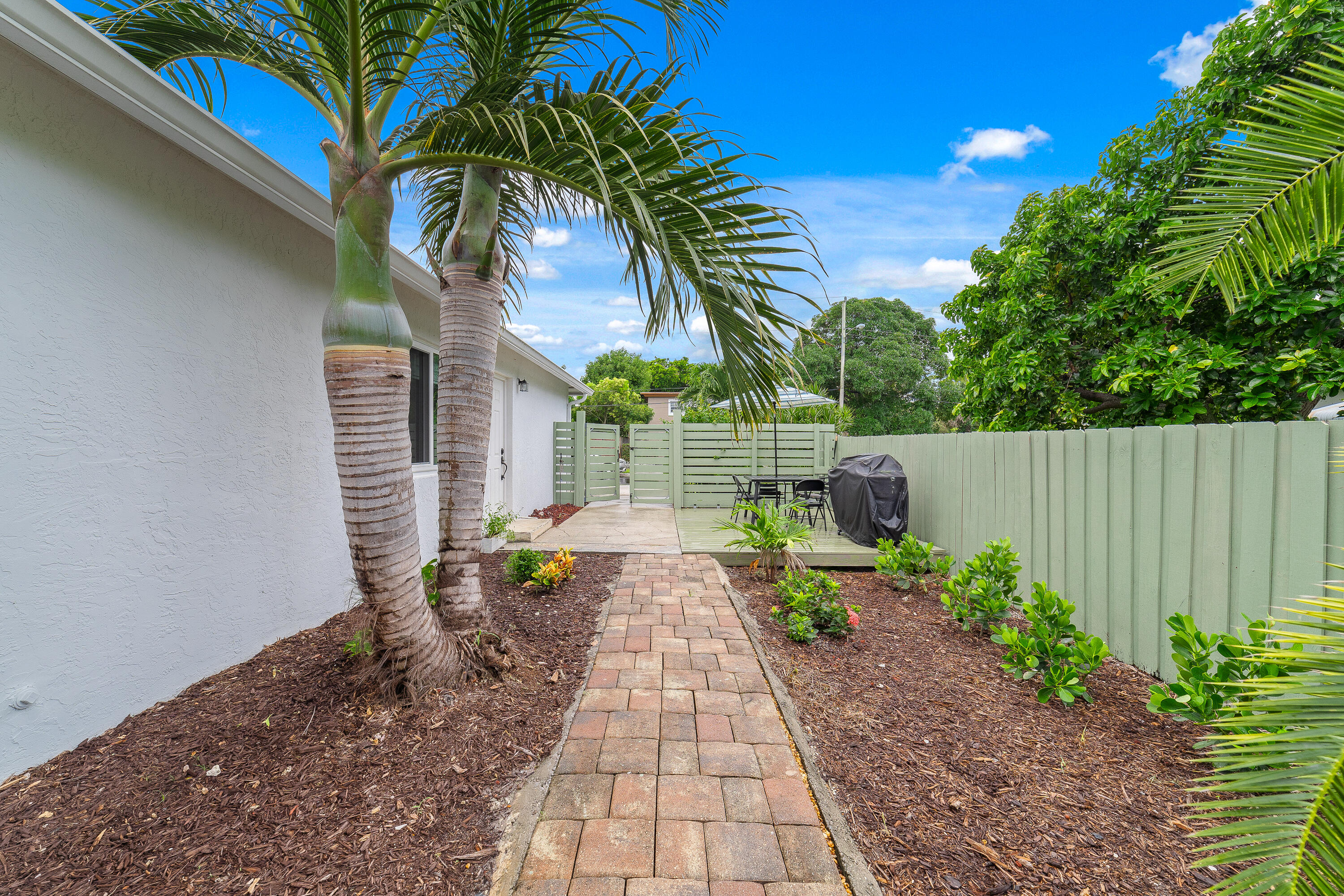 Fenced backyard patio with a brick walkway, palm trees, dining set, and covered grill.