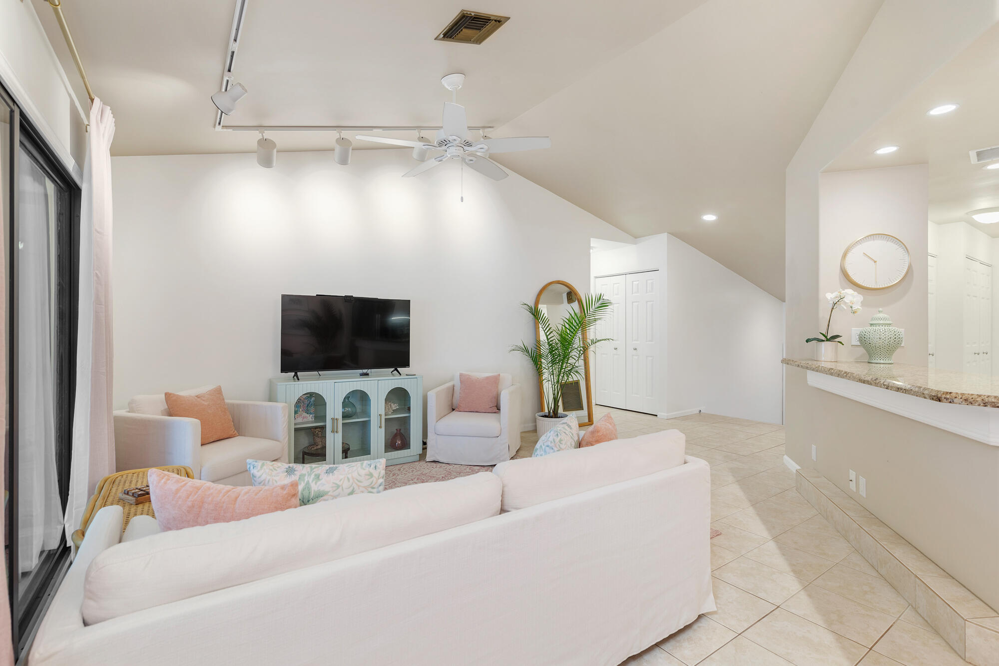 Furnished living room with pale sofas, peach pillows, a vaulted ceiling, and sliding glass doors.