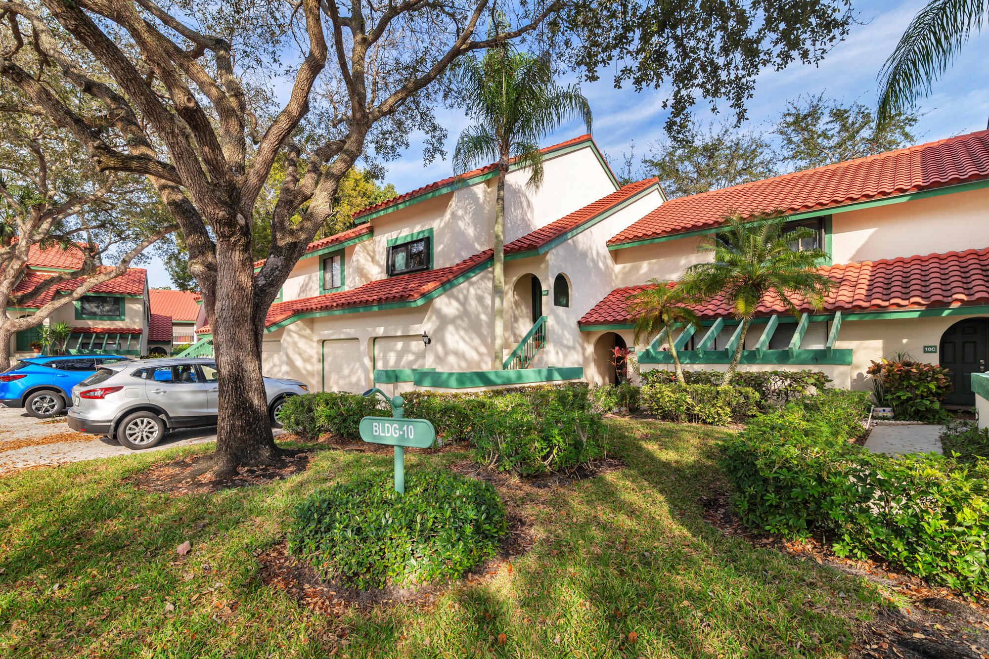 Cream-colored residence with red tile roofs and green trim, framed by a large oak and palms.