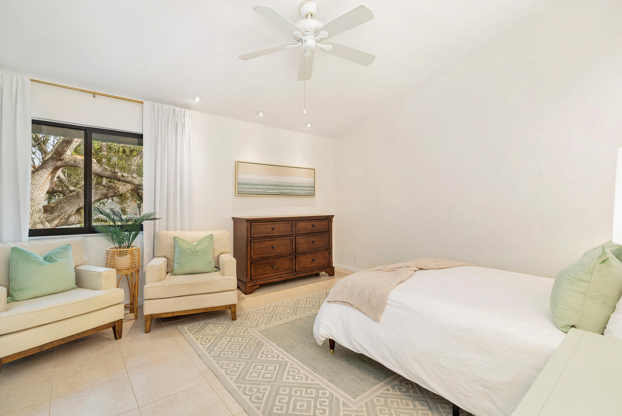 Furnished bedroom with a white bed, cream armchairs, dark wood dresser, and a large window overlooking a tree.