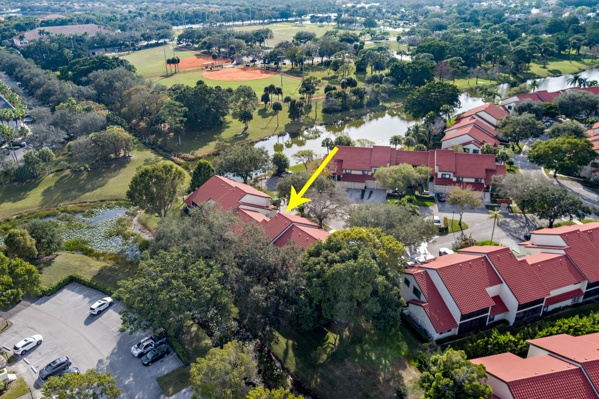 Aerial view of a red-tile-roof condominium marked by a yellow arrow, beside a pond and baseball fields.