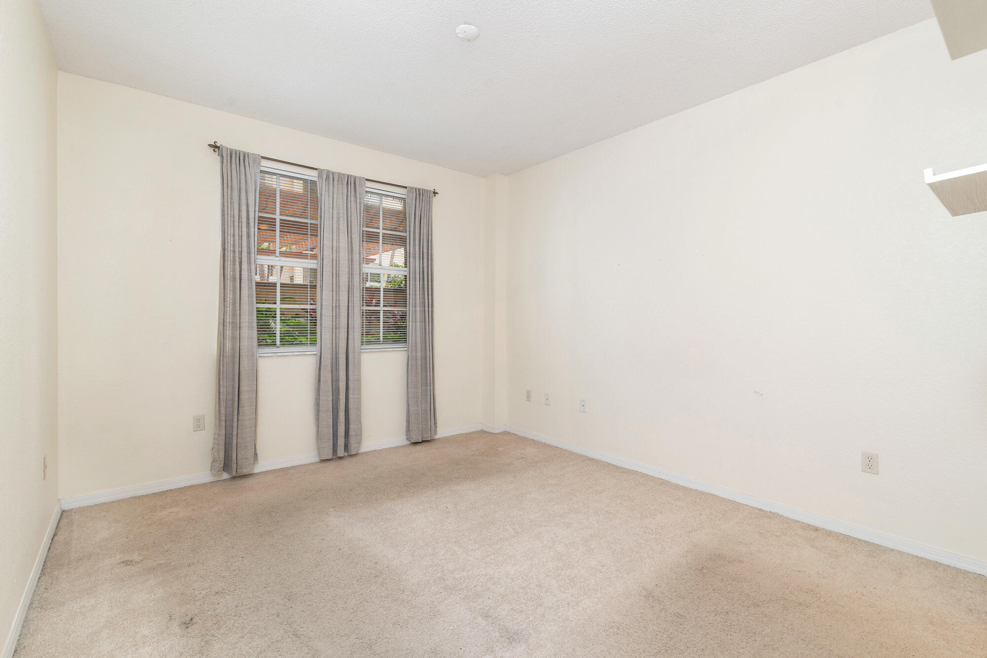 Empty bedroom with beige carpet and two curtained windows with blinds.
