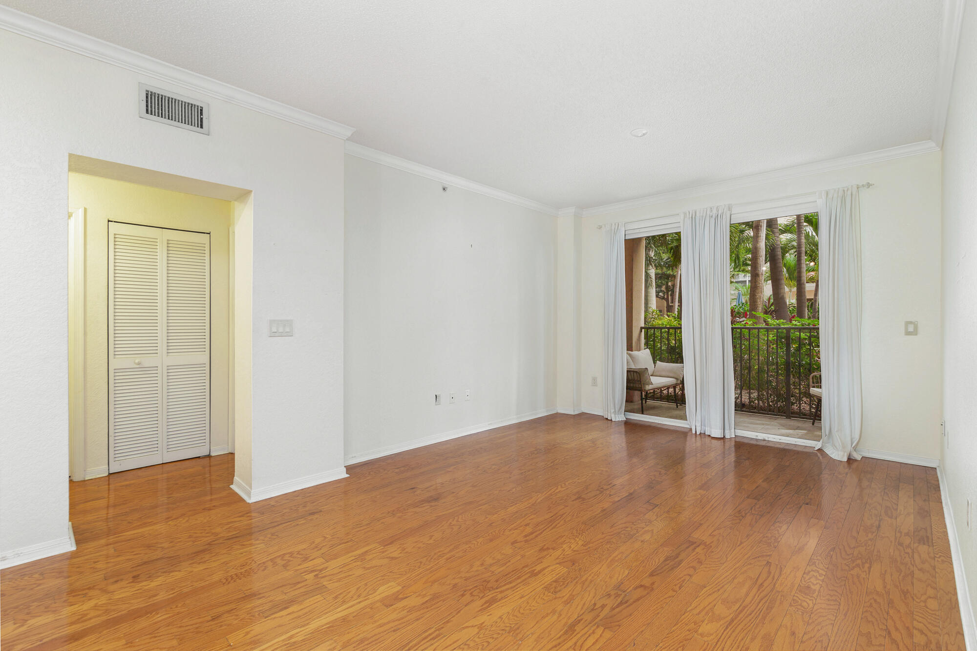 Empty living room with polished hardwood floors and balcony doors overlooking tropical greenery.