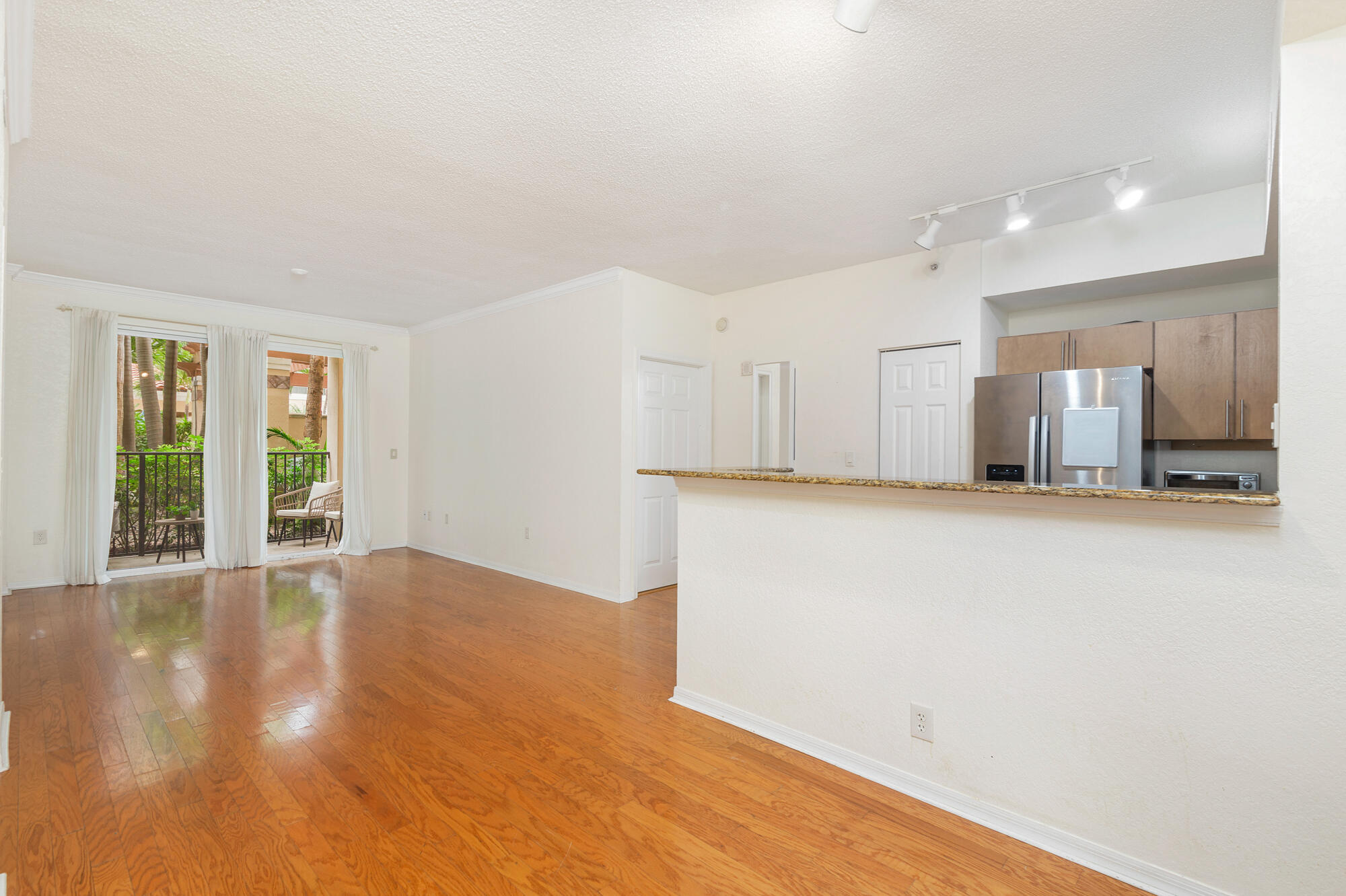 Vacant condo living area with hardwood floors, balcony doors, and a pass-through kitchen.