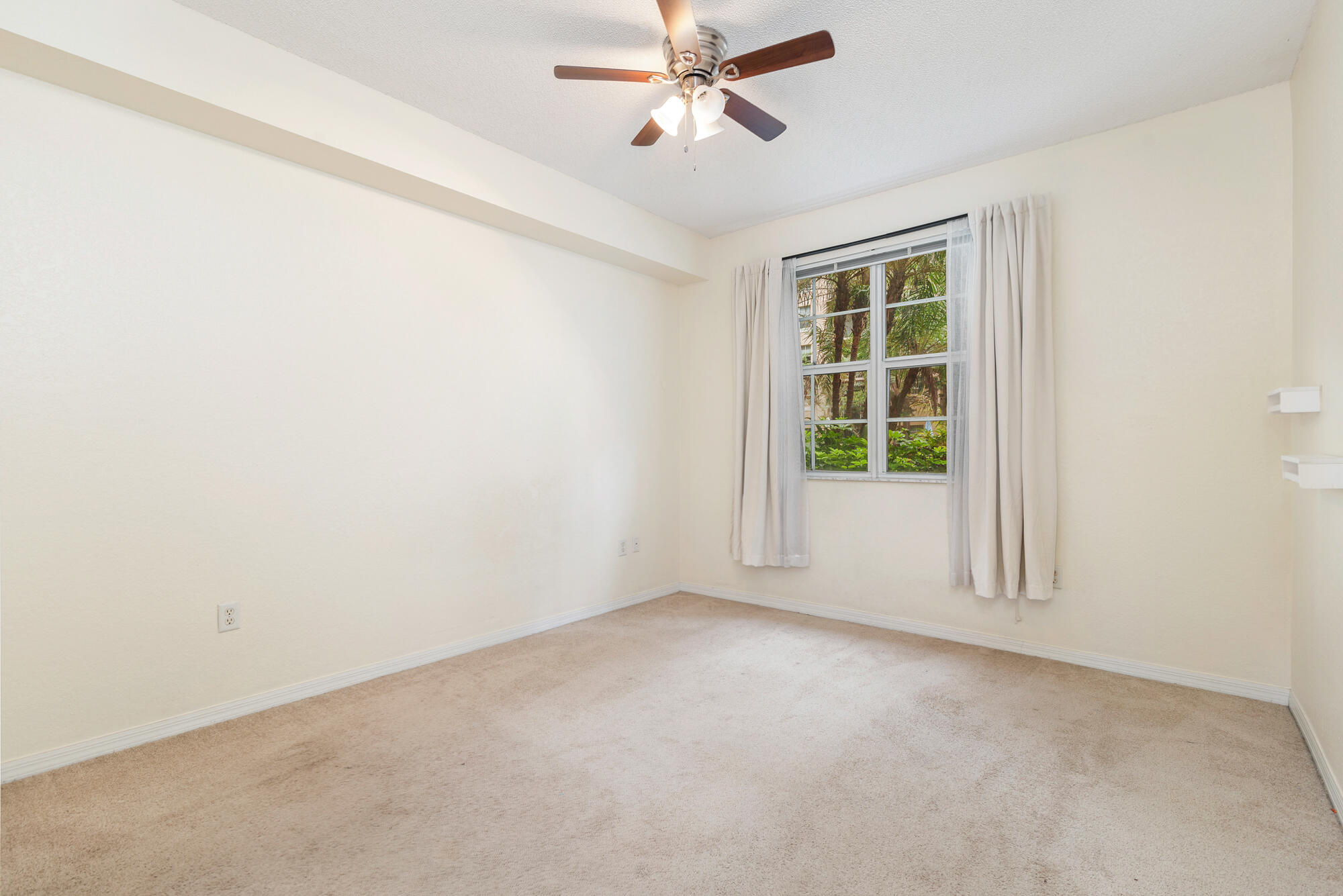 Empty bedroom with beige carpet, a ceiling fan, and a curtained window overlooking greenery.