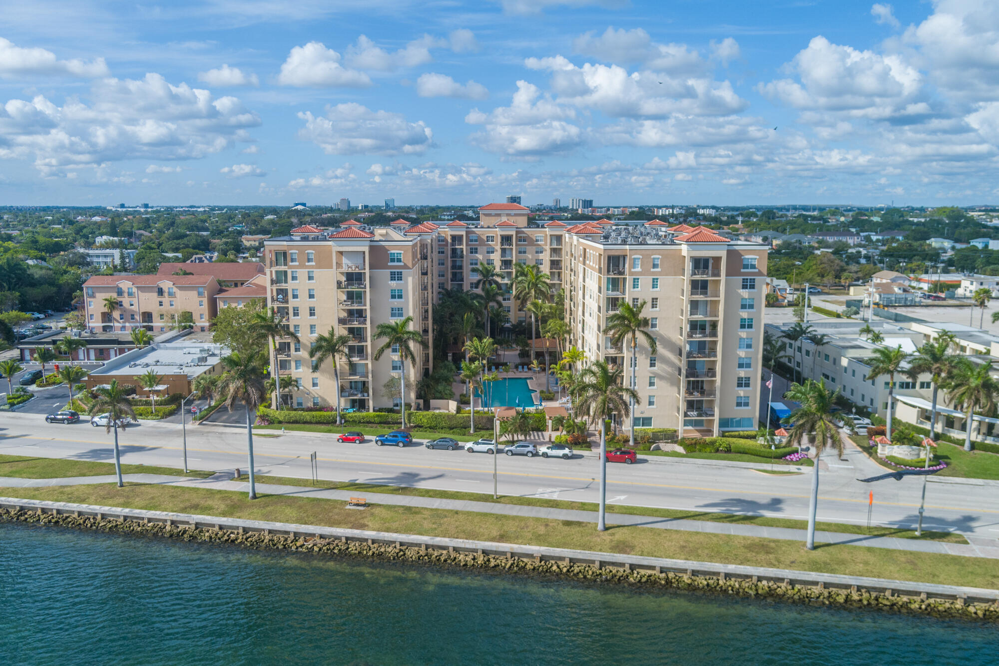 Waterfront condominium with palm trees and courtyard pool, viewed across the road from above.