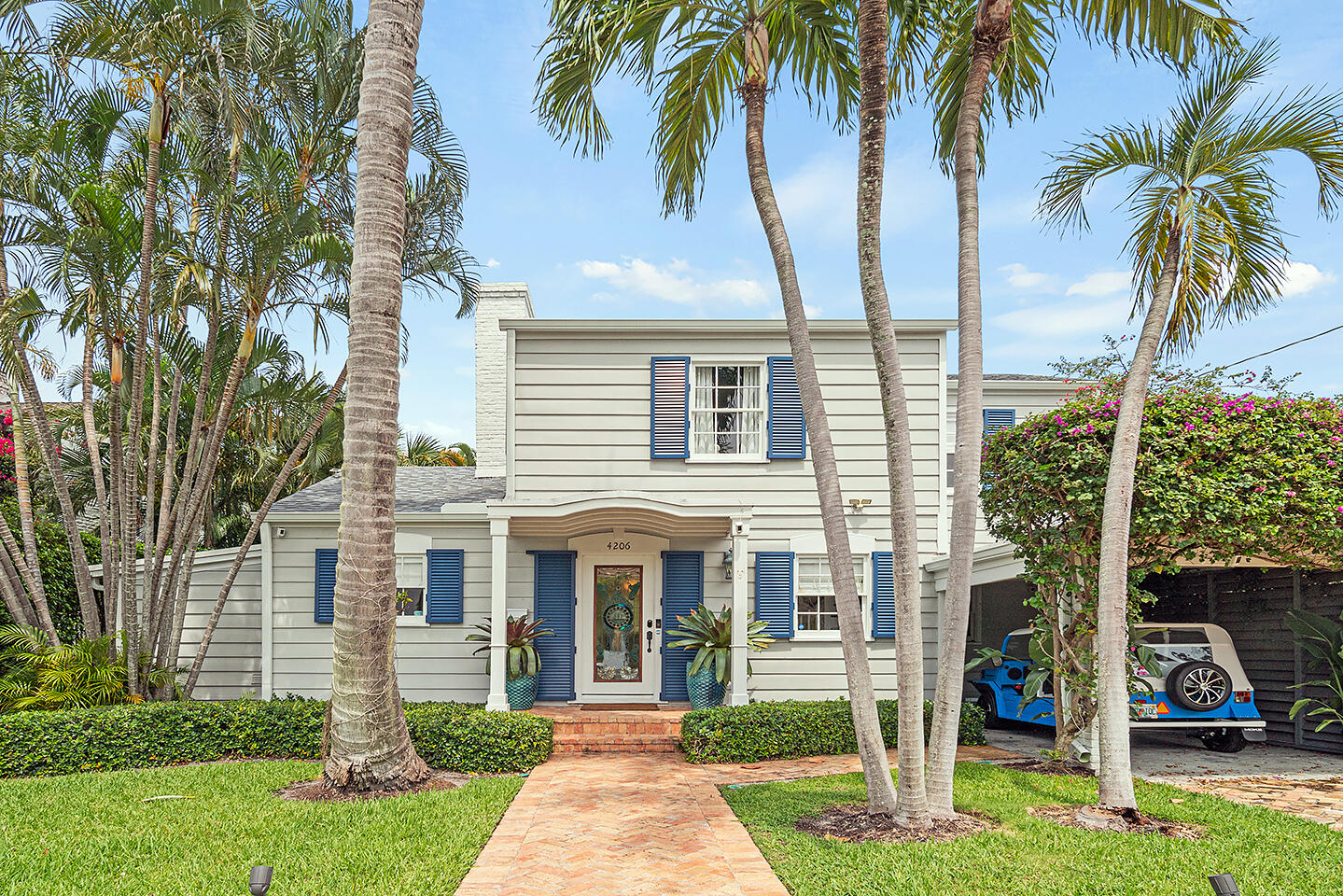 Two-story house with blue shutters, framed by palms and a brick walkway.