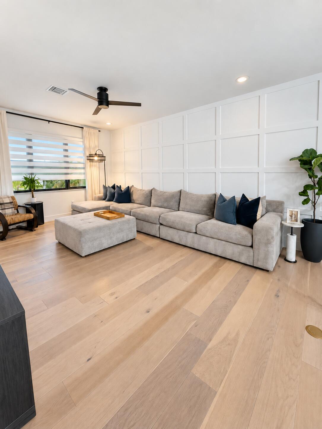 Staged living room with a gray sectional, paneled white wall, and pale wood floors.