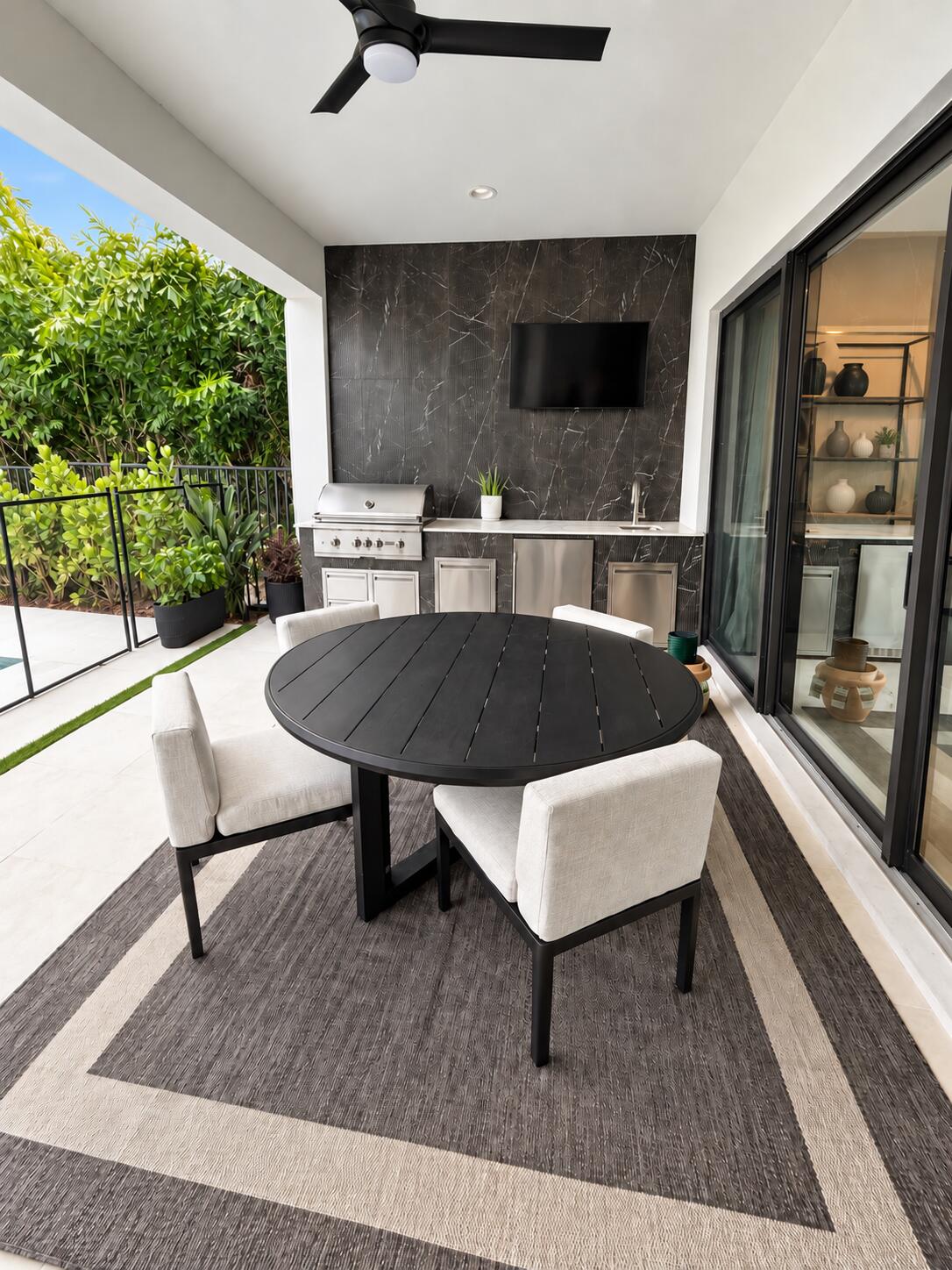 Covered patio with round dining table, outdoor kitchen, and lush greenery beyond the railing.
