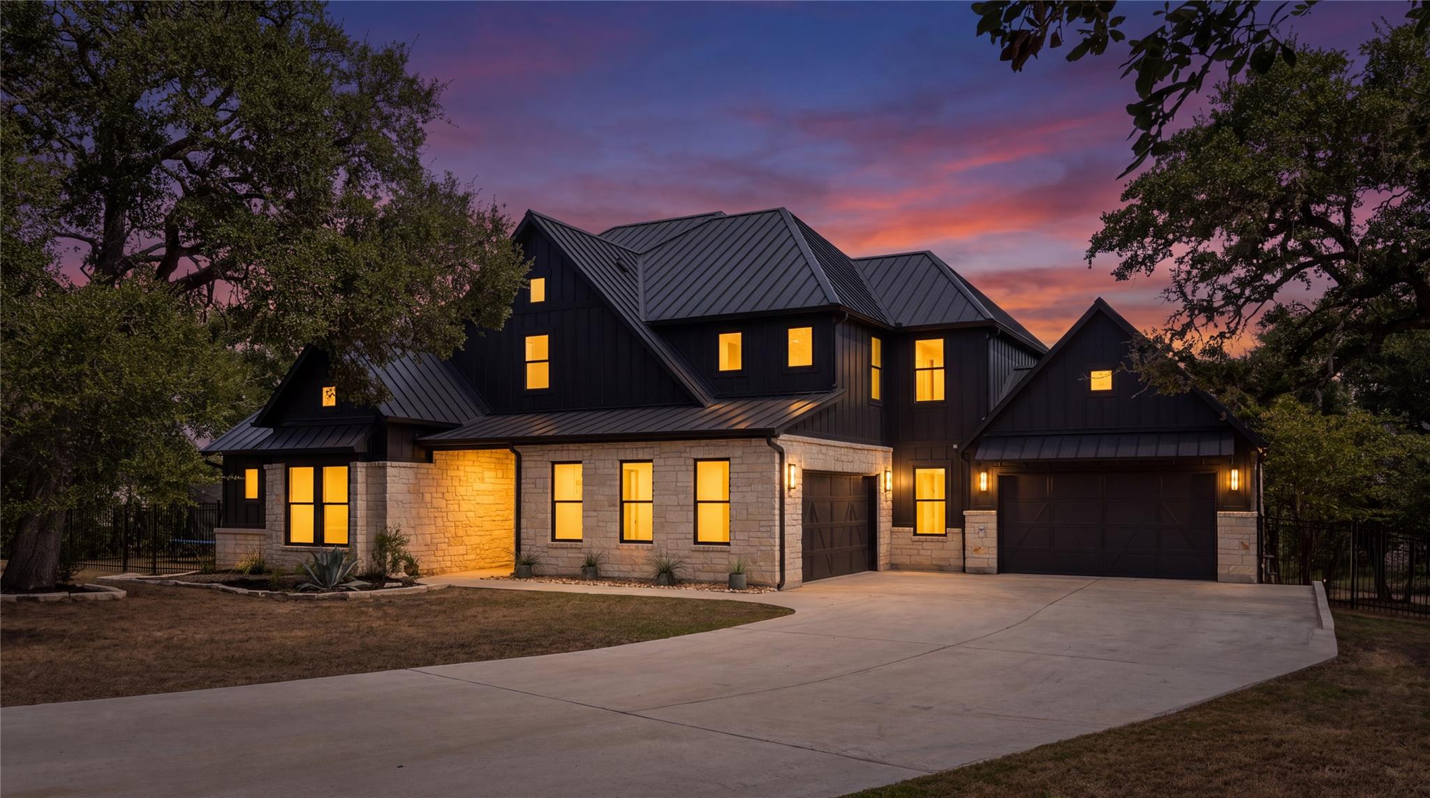 Two-story stone and dark-sided home with glowing windows, steep metal roofs, and a broad driveway at dusk.