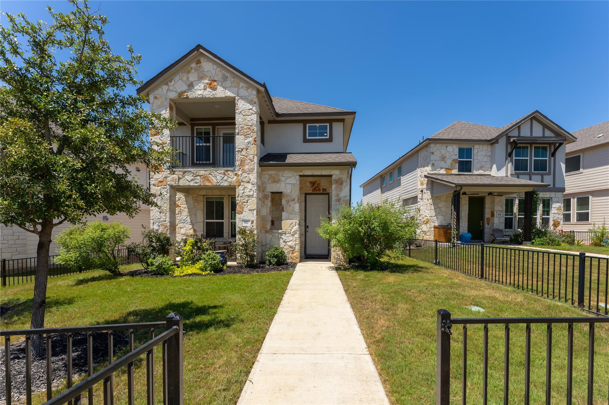 Two-story stone-front home with an upper balcony, landscaped lawn, and walkway through a black metal gate.