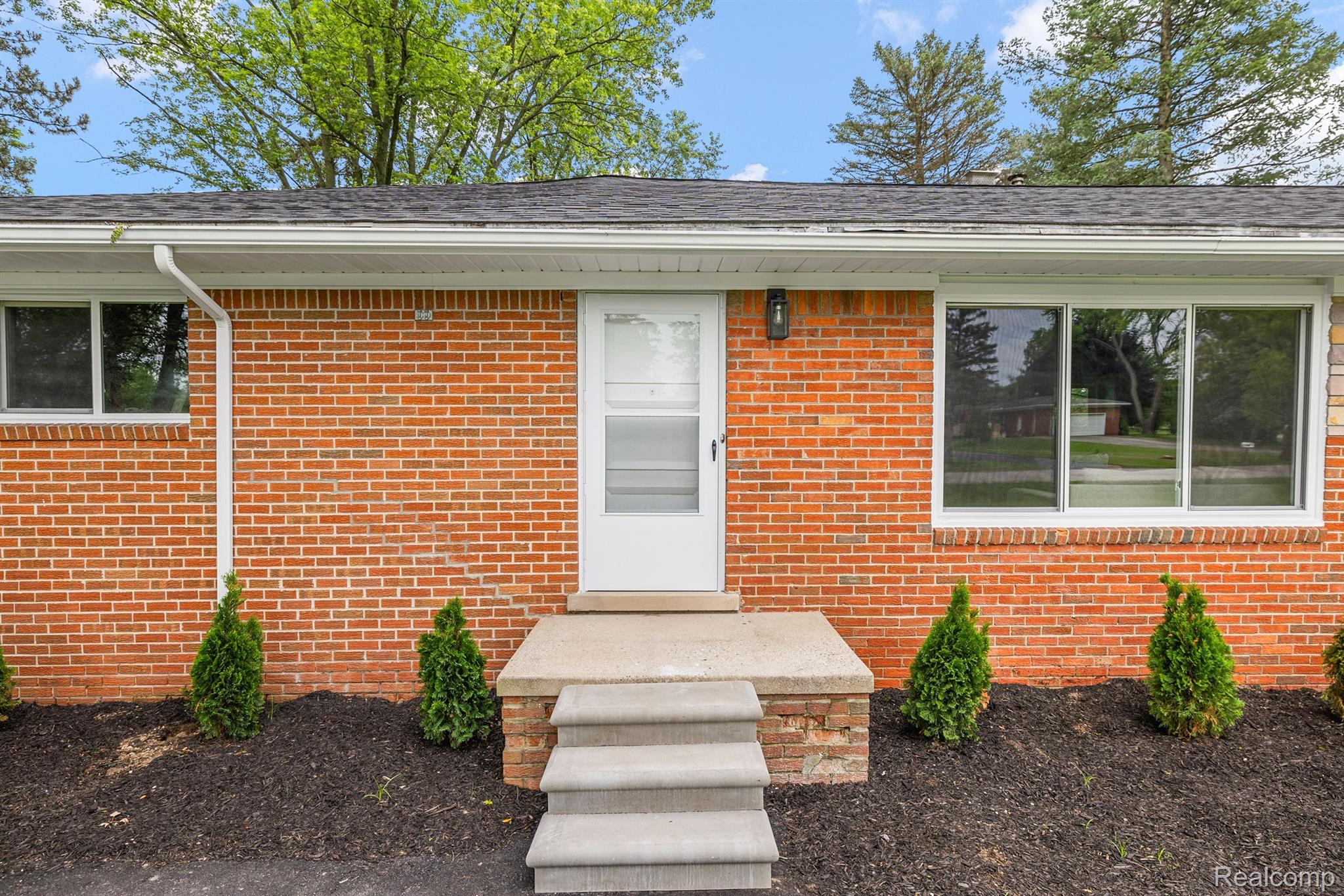 Red-brick home entrance with white door, concrete steps, and evergreen shrubs.