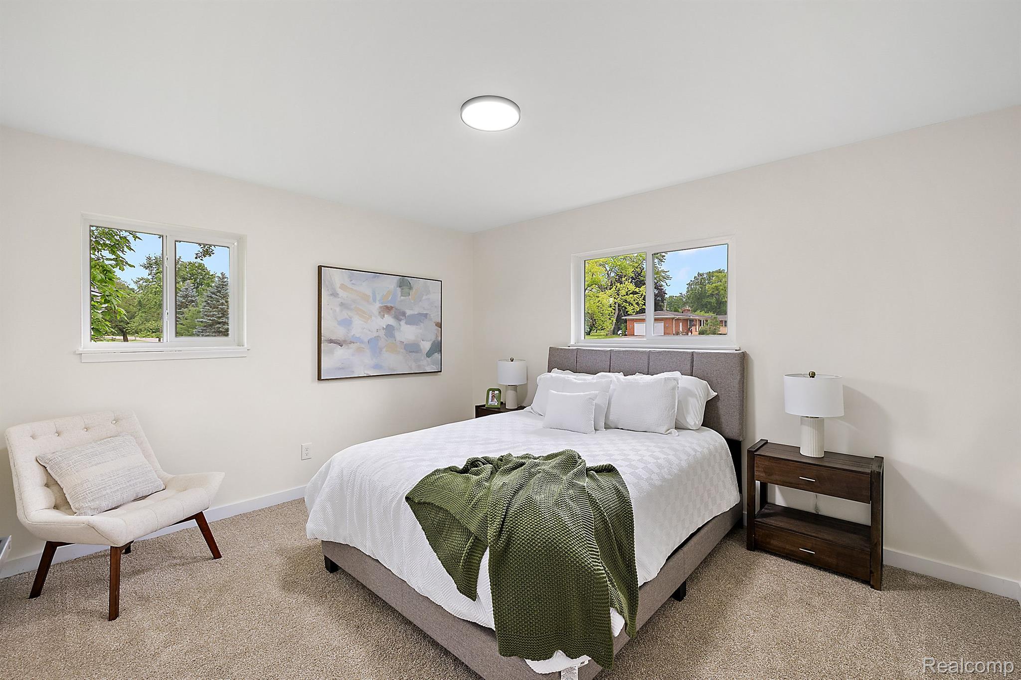 Staged bedroom with a white bed, green throw, gray headboard, and windows overlooking trees.