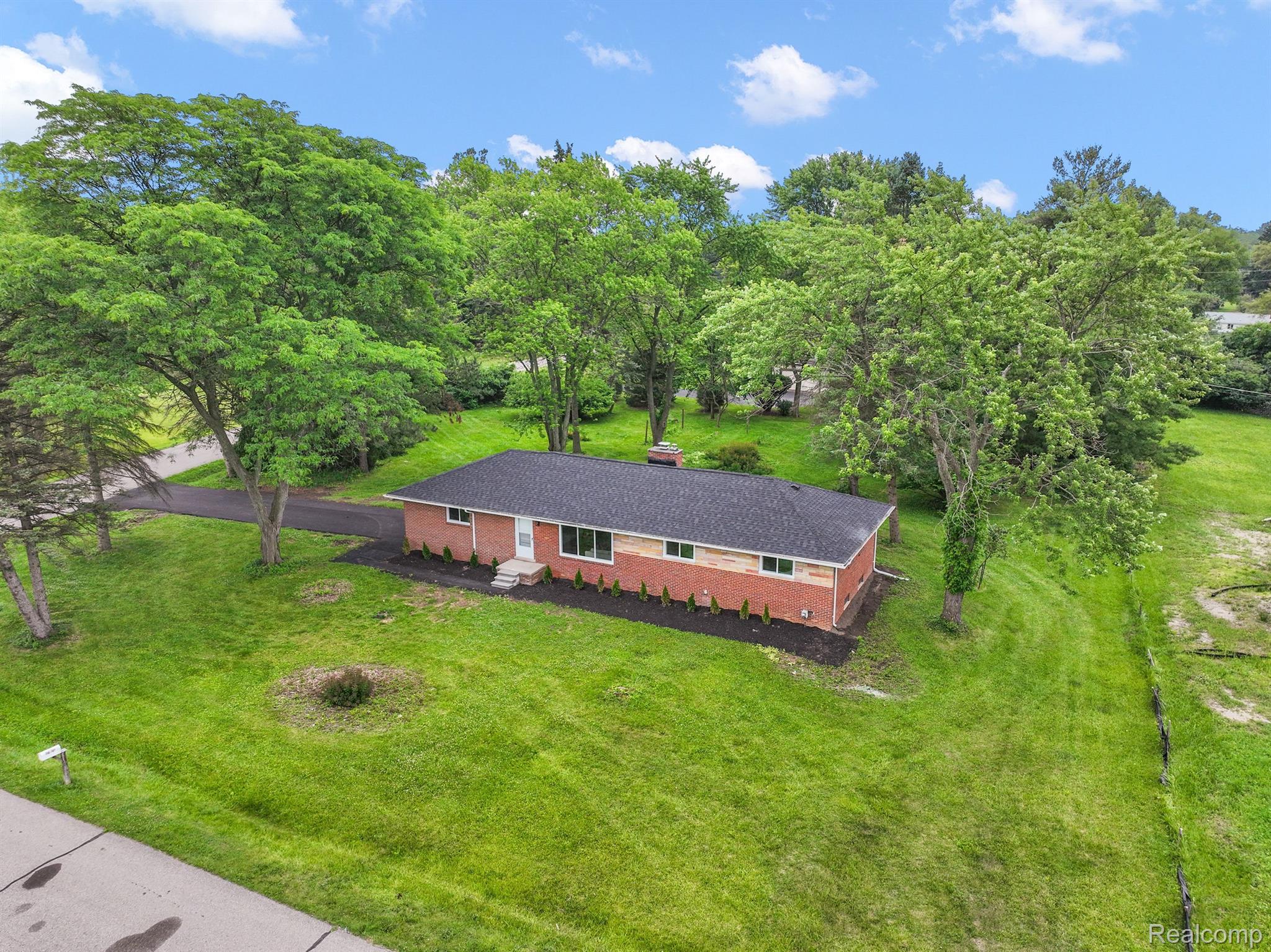Aerial view of a red-brick ranch house on a spacious green lot shaded by mature trees.
